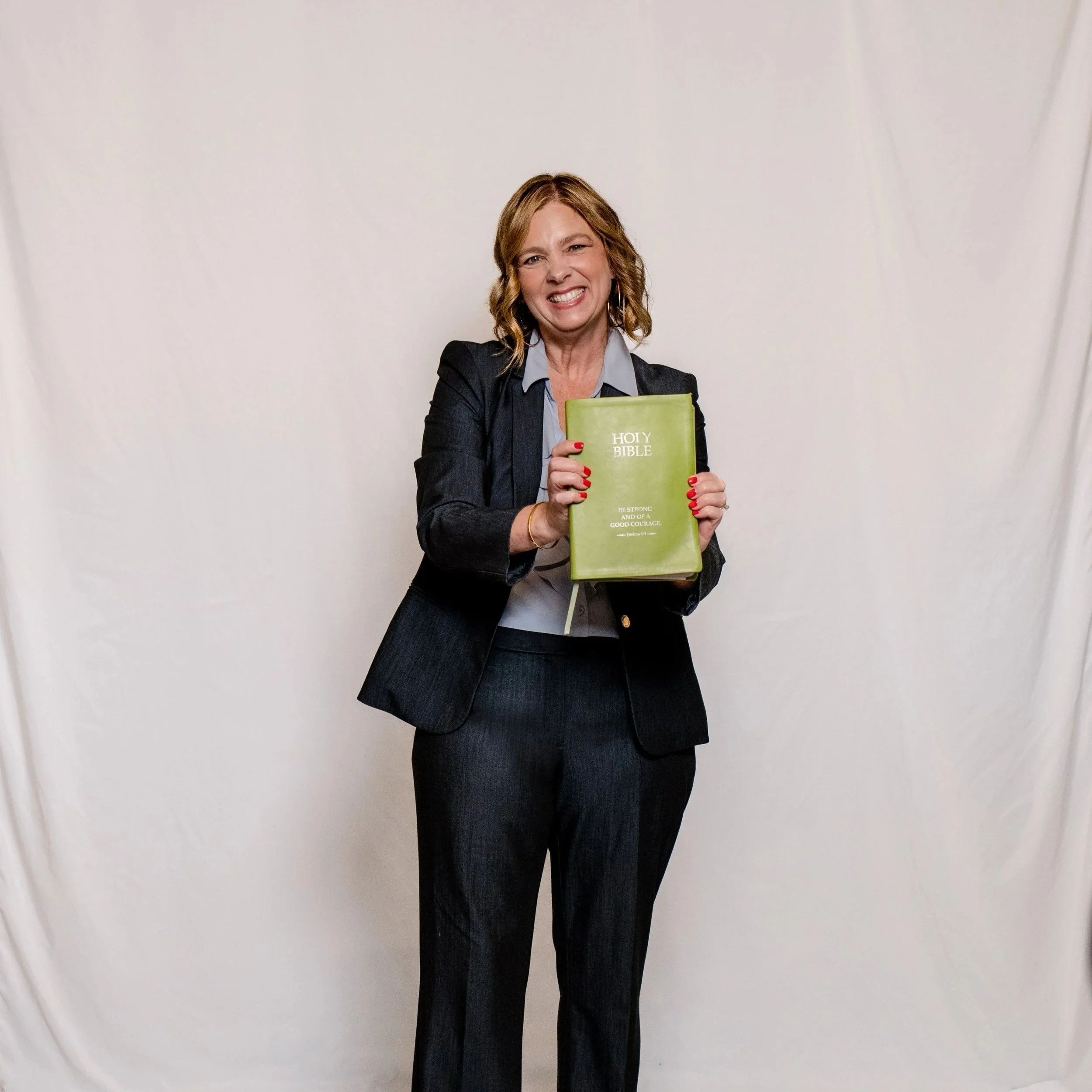 A woman with shoulder-length wavy hair smiling and squinting, holding a green Holy Bible with a positive message on the cover. She is dressed in a dark blazer and light shirt, standing against a plain white background.