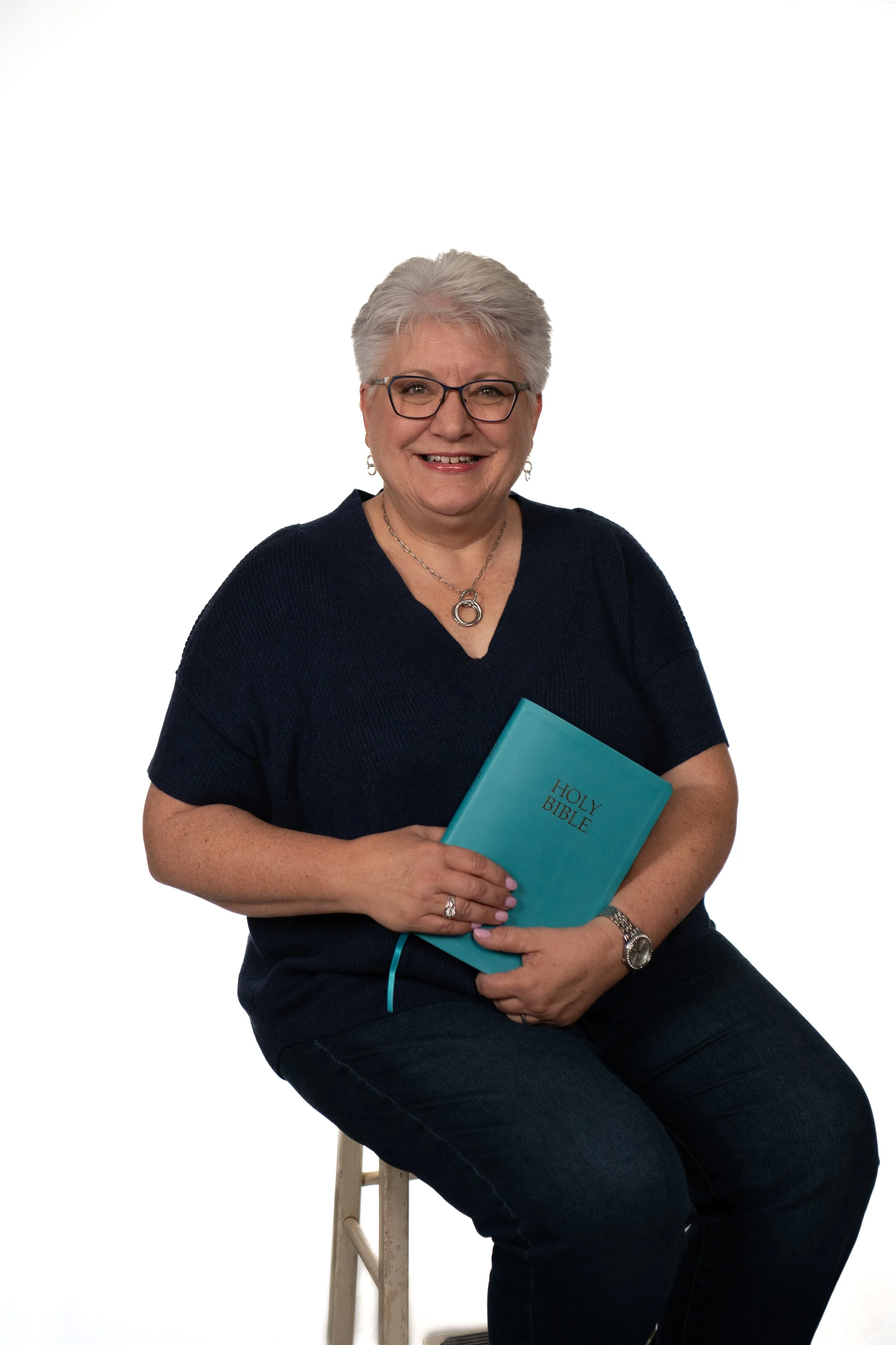 A smiling older woman with short gray hair, glasses, and jewelry, wearing a dark blue top and jeans, sitting on a wooden stool, holding a blue Holy Bible, against a white background.