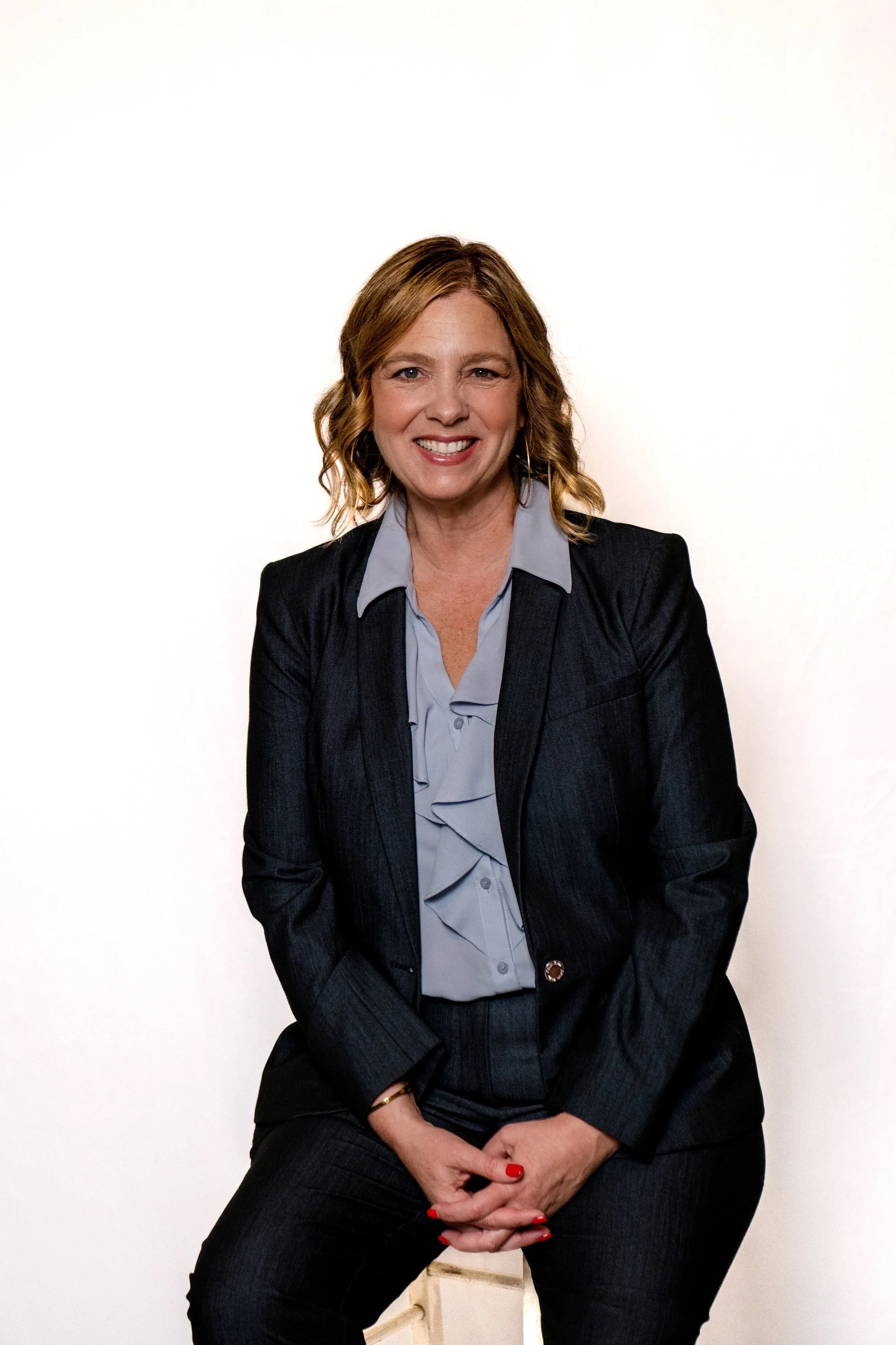 A woman with wavy, shoulder-length blonde hair, smiling, wearing a dark blazer over a light blue shirt, sitting against a plain white wall.