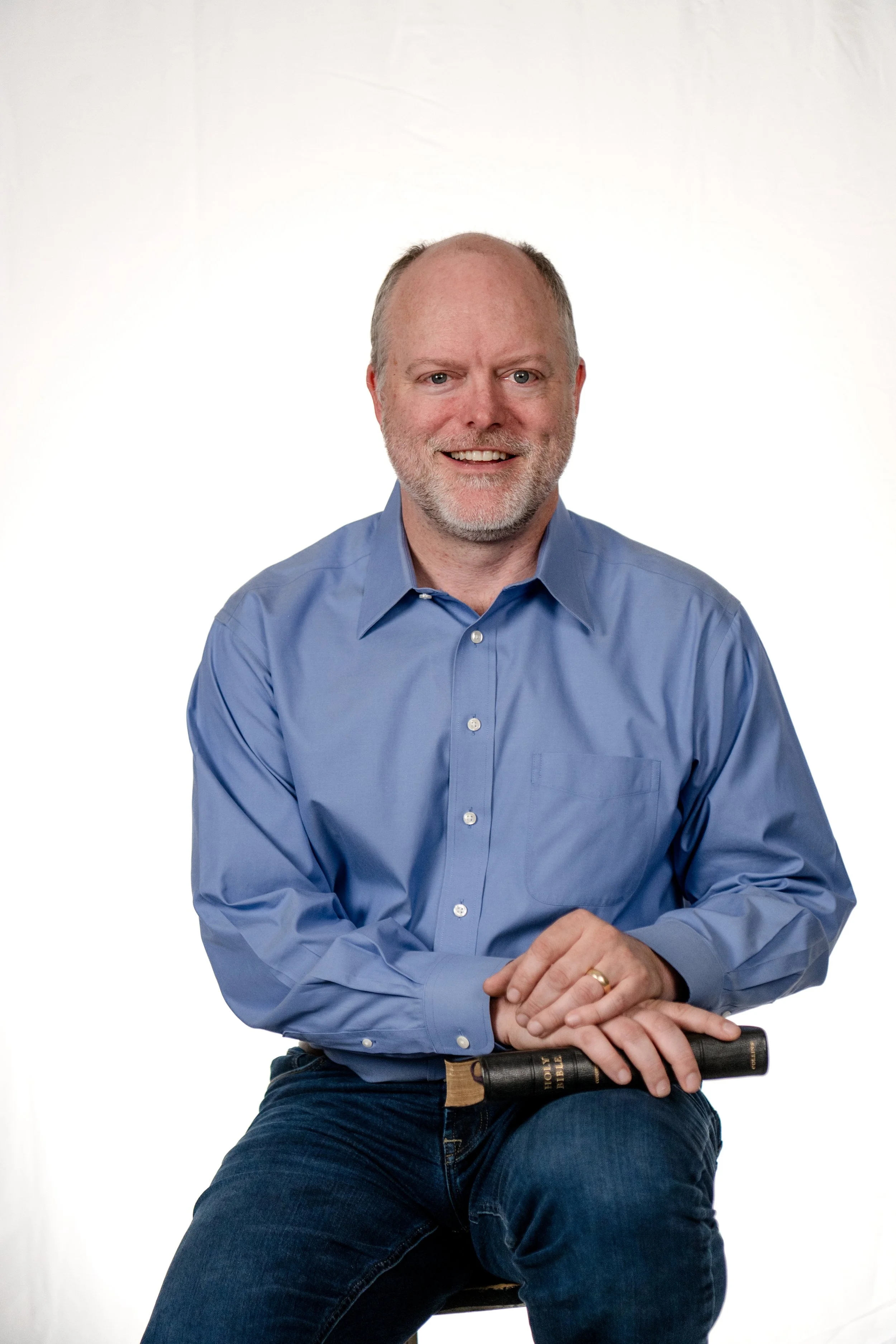 A middle-aged man with a beard, wearing a blue dress shirt and jeans, sitting against a plain white background. He is holding a Bible in one hand and has his other hand resting over it. He is smiling and looking directly at the camera.