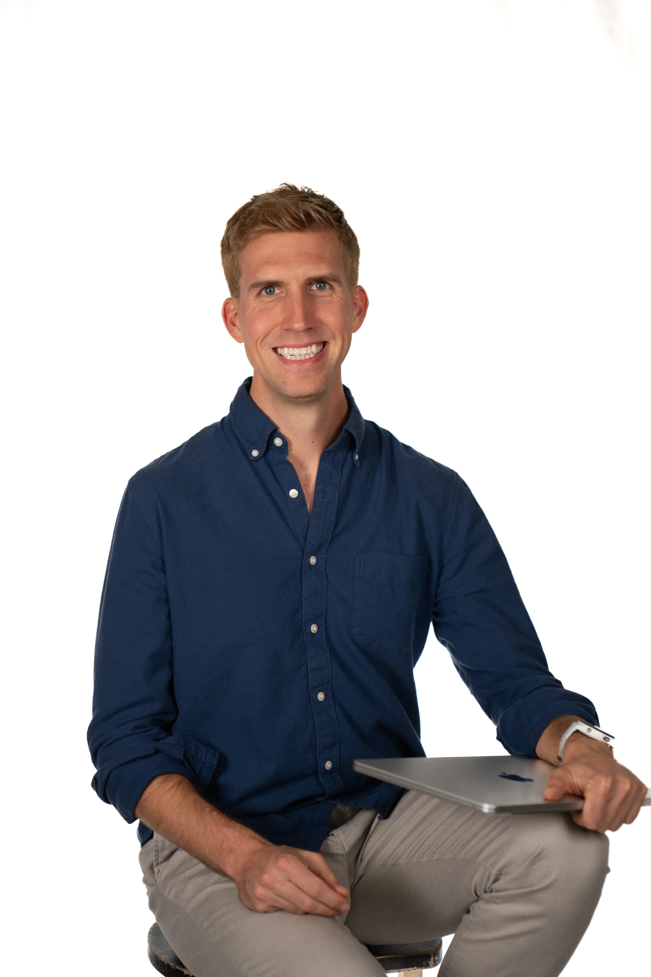 A young man with light brown hair, wearing a dark blue button-up shirt, sitting on a stool and holding a closed silver laptop, smiling at the camera against a white background.