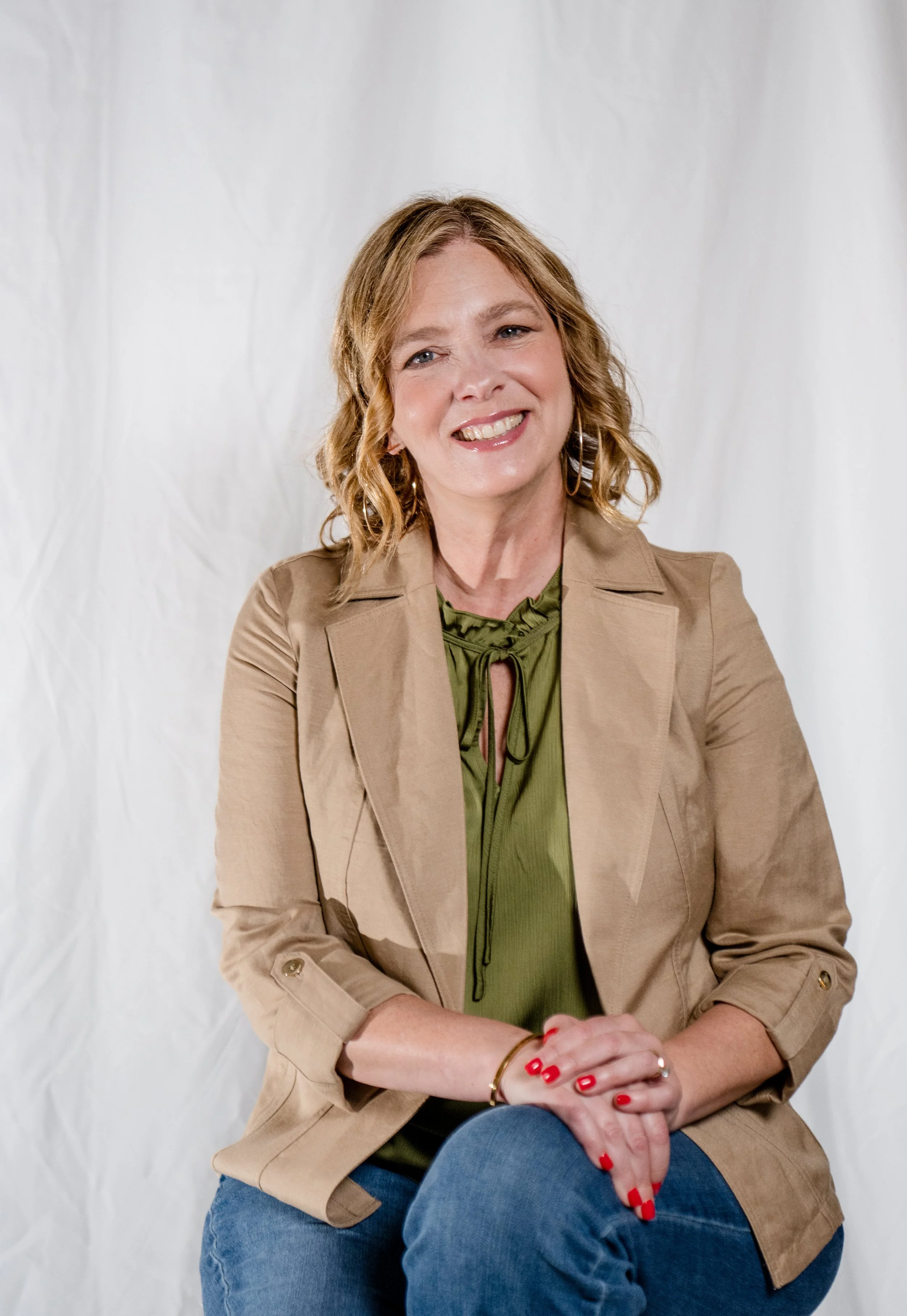A smiling woman with shoulder-length wavy blonde hair, wearing a beige blazer over a green top, and sitting with her hands clasped in her lap against a plain white background.