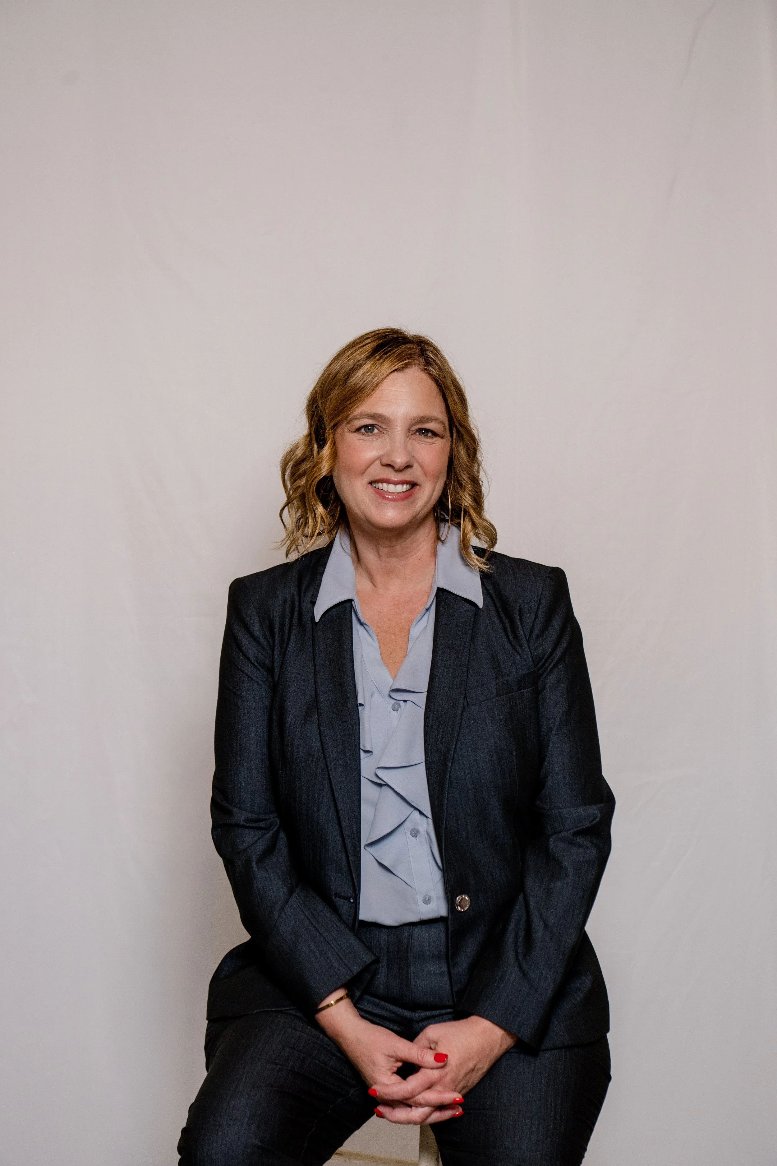 A woman with shoulder-length wavy blonde hair and light skin, smiling, wearing a dark blazer over a light blue shirt, seated against a plain light-colored wall.