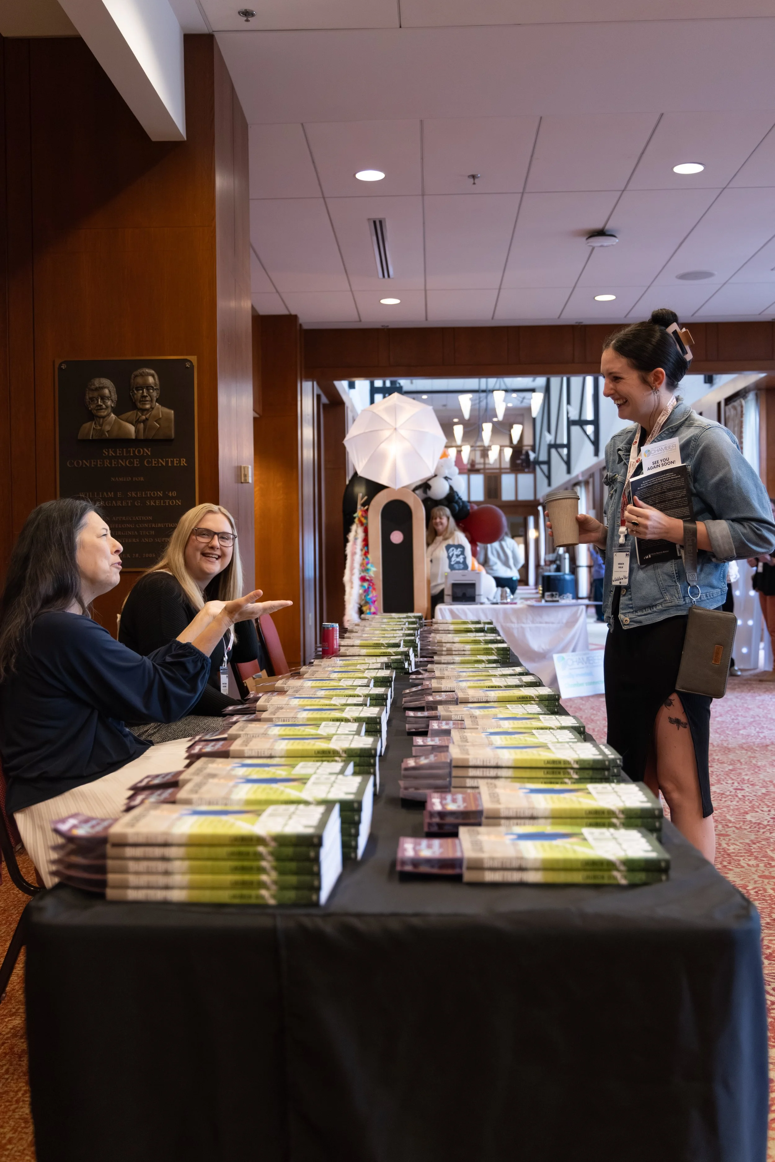 A woman with short dark hair, wearing a denim jacket and a name badge, is smiling and holding a cup of coffee while talking with two women seated at a table covered with stacks of books. The table is inside a conference center or hotel ballroom, with a wooden wall and a plaque in the background. Other attendees and decorations, including an umbrella and plush panda toys, are visible in the background.