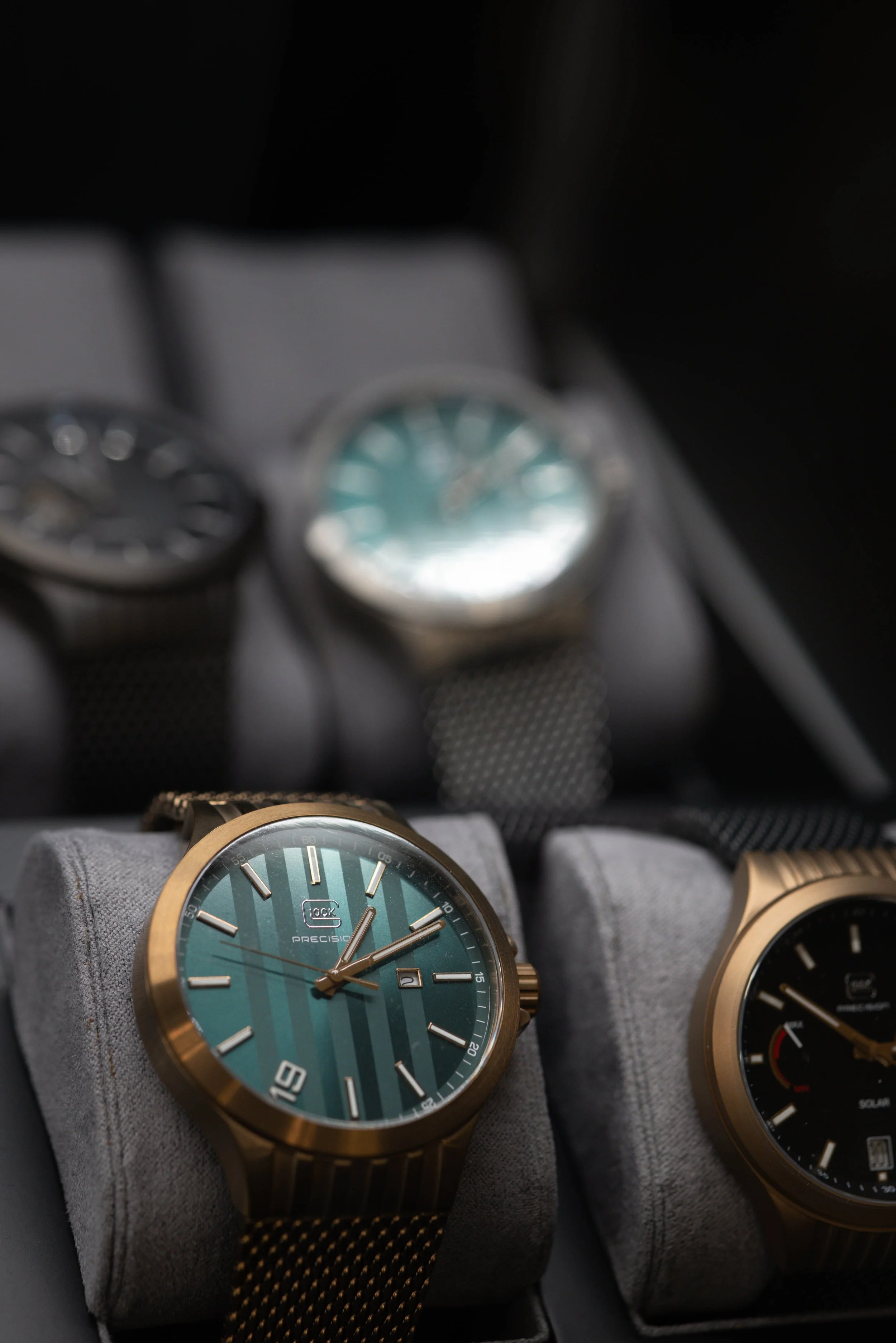 Several wristwatches with metallic bands and round faces placed on a soft gray watch holder, with a blurred background of more watches.