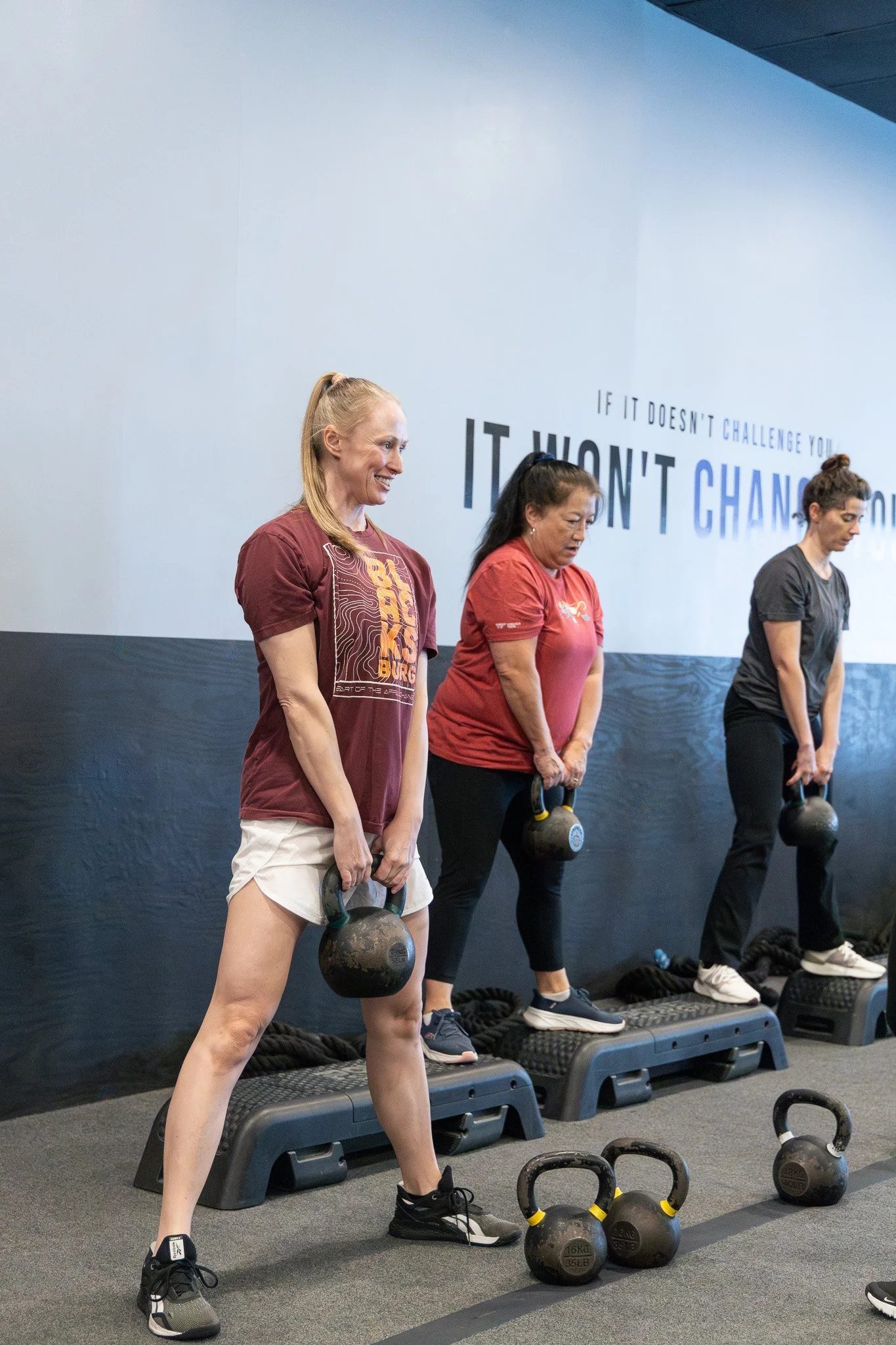 Women in a gym, standing on step platforms, holding kettlebells, participating in a fitness class.