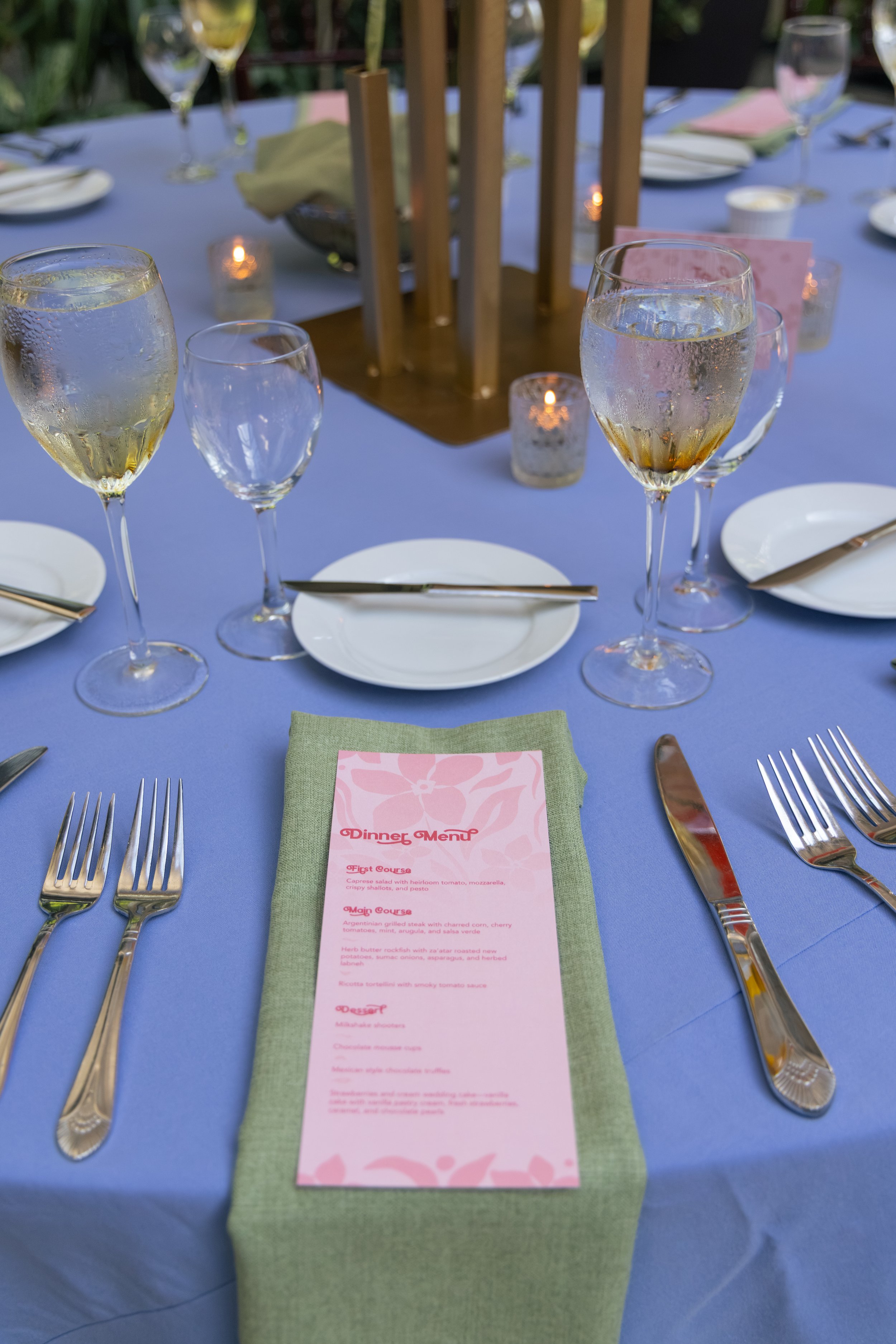 A round table with a lavender tablecloth set with white plates, silverware, and glasses of sparkling white wine, decorated with a wooden centerpiece and small candles, and a pink dinner menu placed on a green napkin in the center.