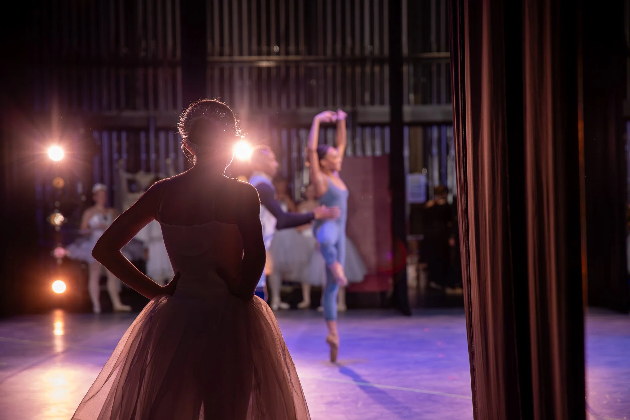 Backstage view of ballet dancers preparing for performance with a young ballerina in the foreground wearing a pink tutu, and other dancers stretching and warming up in the background with stage lights shining.