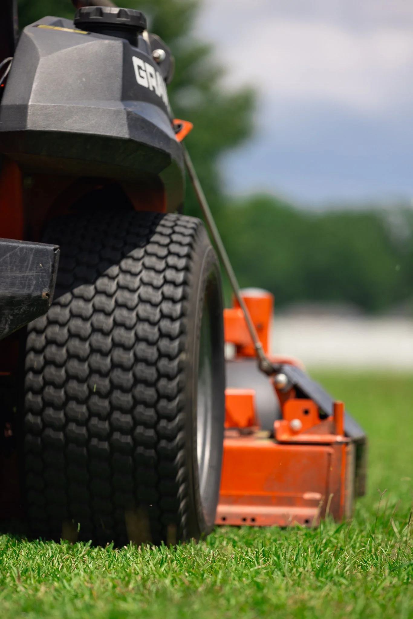 Close-up of a lawn mowing machine's rubber tire with a patterned tread, orange equipment in the background, and a grassy lawn.
