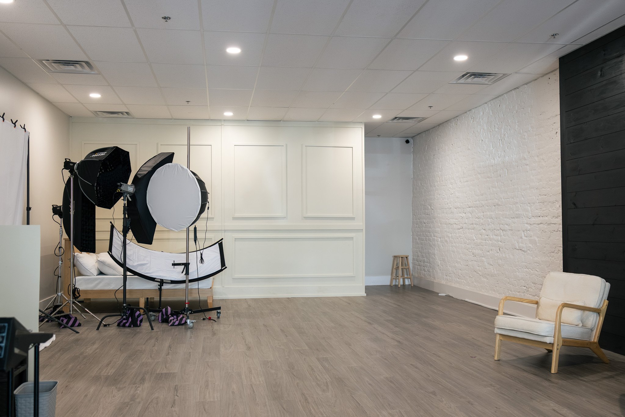 Photography studio with lighting equipment and a white backdrop in a room with white walls, a black wall panel, wooden flooring, and a white armchair.