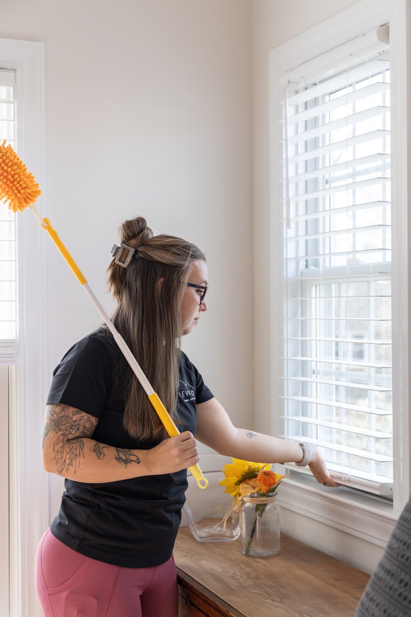 A woman with tattoos on her left arm and glasses, wearing a black t-shirt and pink leggings, is standing by a window with white blinds in a room with cream-colored walls and a wooden table. She is holding a yellow cleaning duster and is reaching to clean the window. On the table, there is a glass jar with yellow and orange flowers and a basket with more flowers inside.