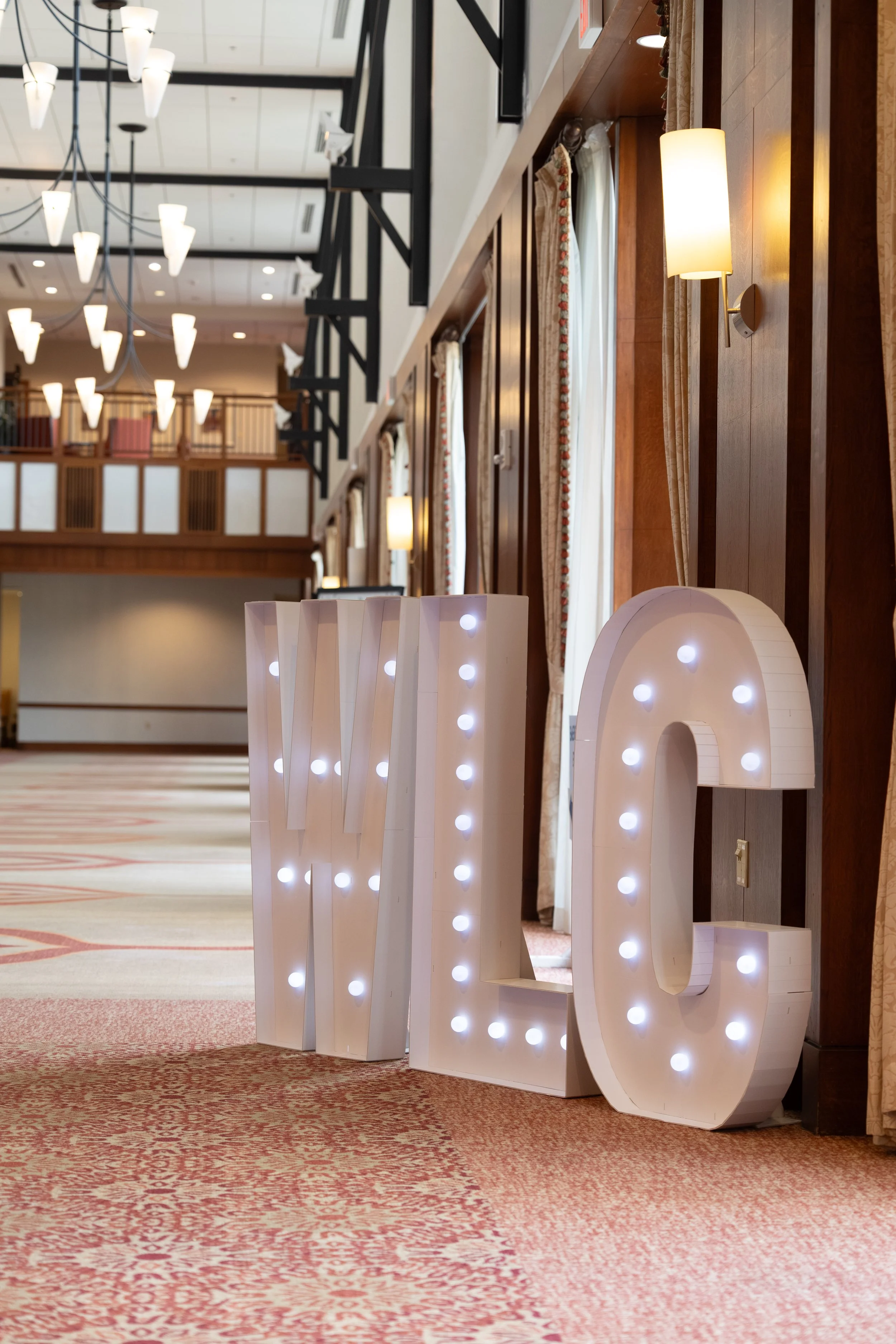 Large illuminated white letter sculpture spelling 'LOVE' placed on a carpeted floor in a spacious, well-lit hotel lobby with wood-paneled walls, elegant curtains, and high ceilings with modern pendant lighting.
