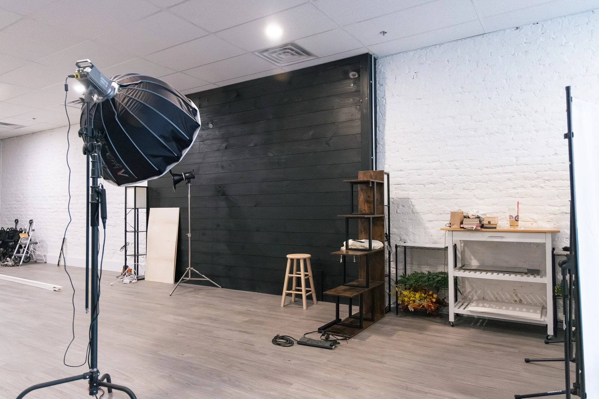 Photography studio or workspace with lighting equipment, a black wooden wall, white brick wall, a stool, shelves with plants, and various items on a white table.