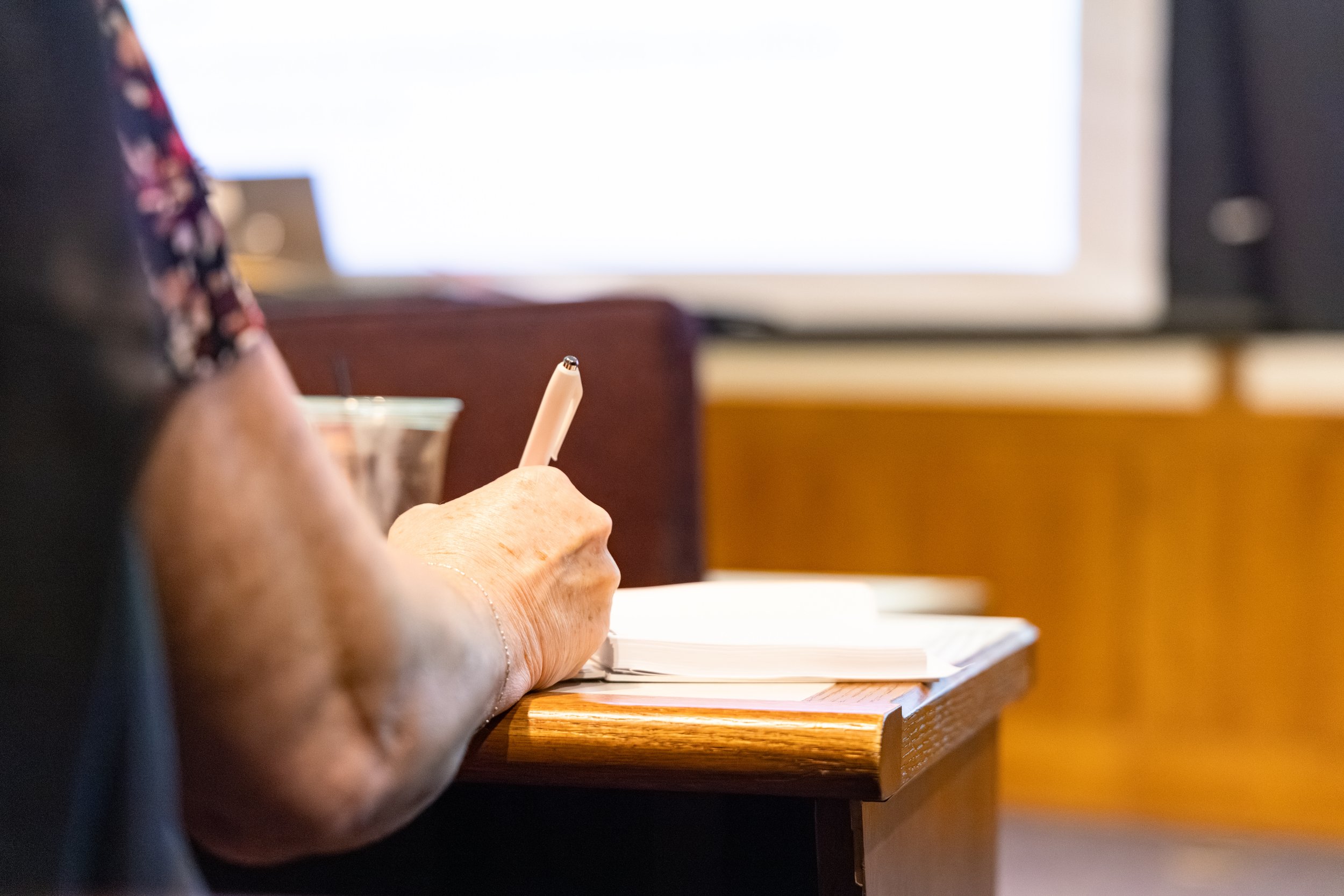 Close-up of an elderly person's hand holding a pen and writing on a piece of paper on a wooden desk, with a large computer monitor in the background.