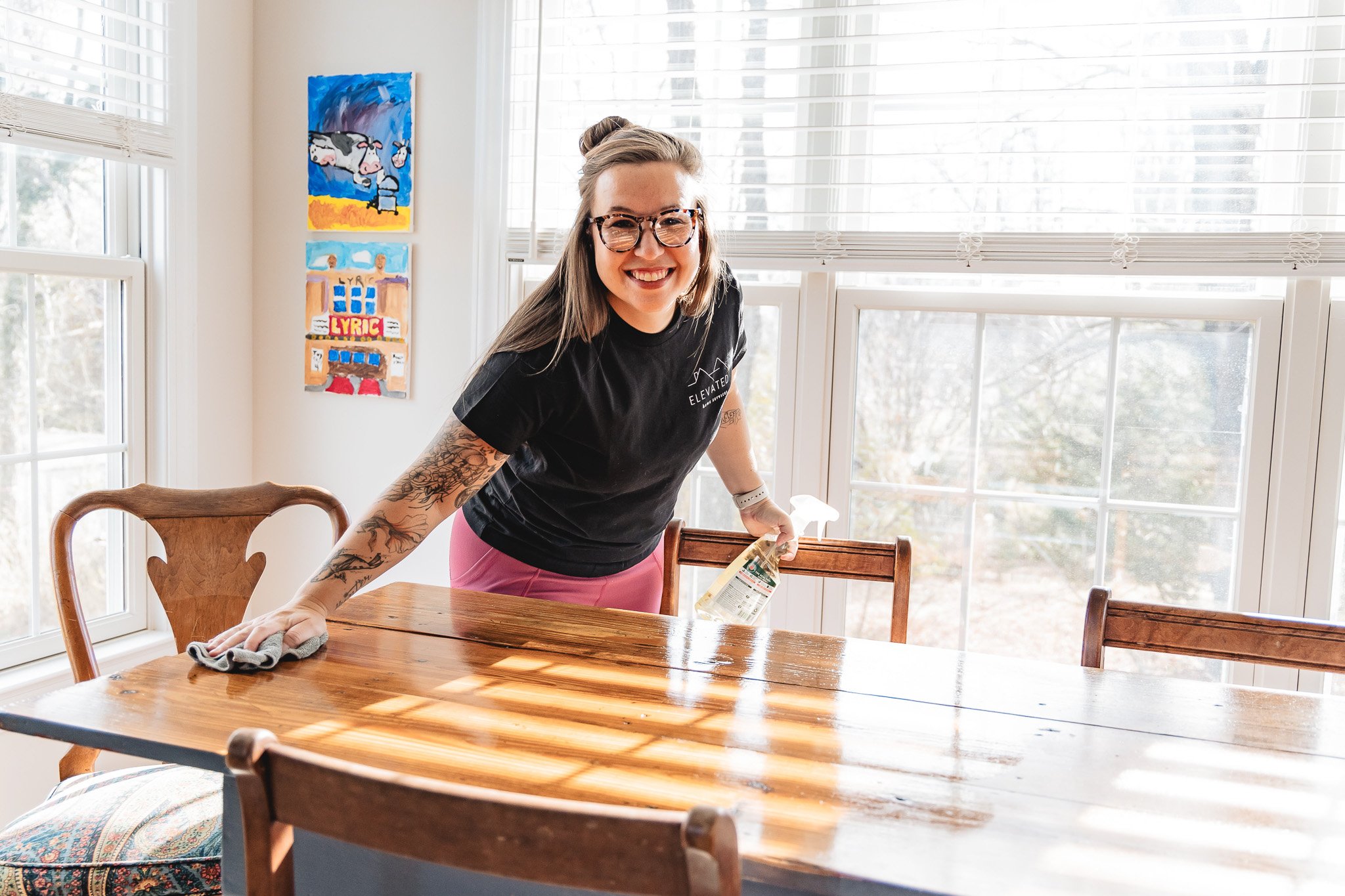 A woman with tattoos, glasses, and a black t-shirt smiling and cleaning a wooden dining table with a cloth and spray bottle of cleaning solution inside a bright room with large windows, art on the wall, and chairs around the table.