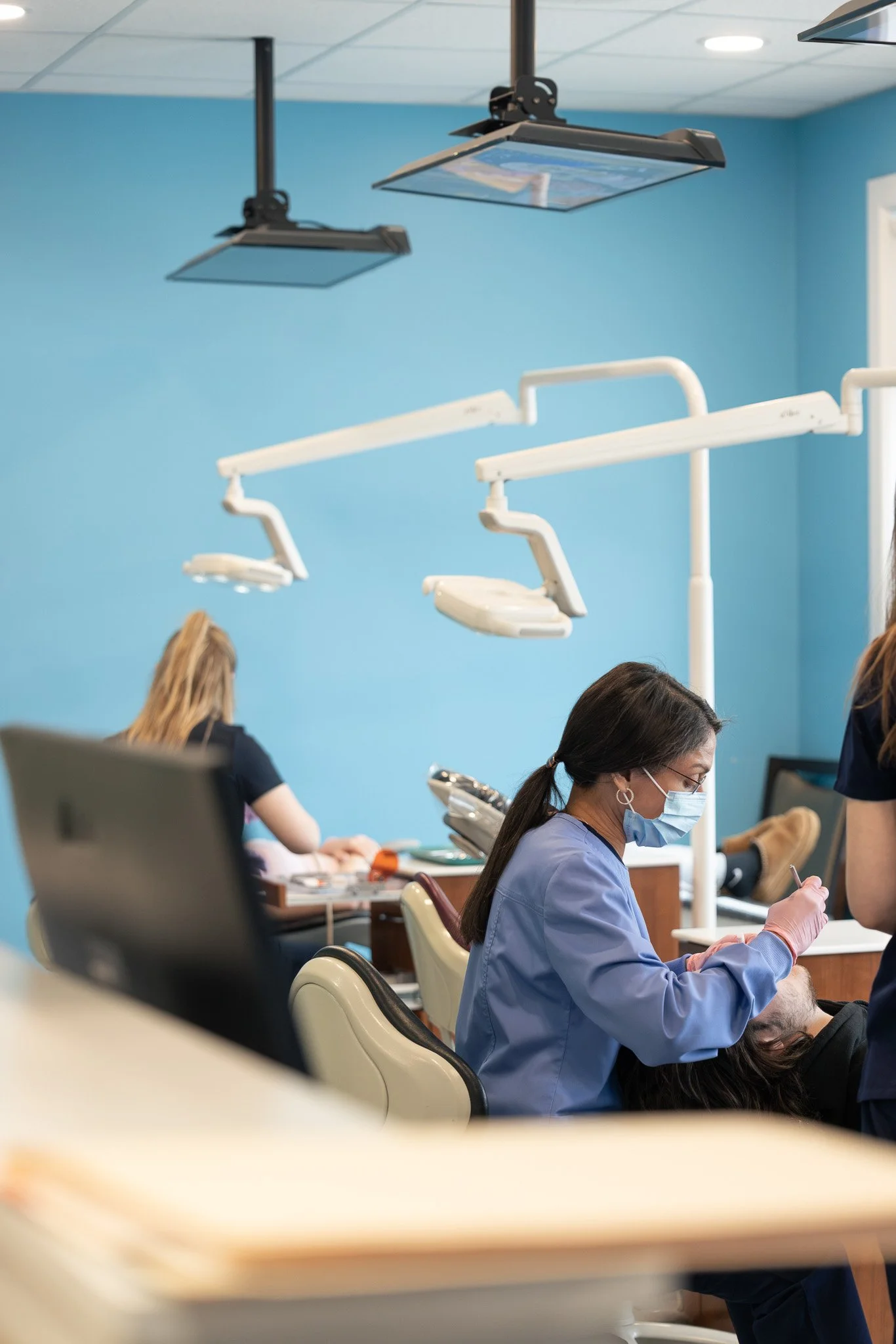 Dental clinic staff members, including a woman in scrubs and face mask, working at dental chairs with overhead lights in a brightly colored room.