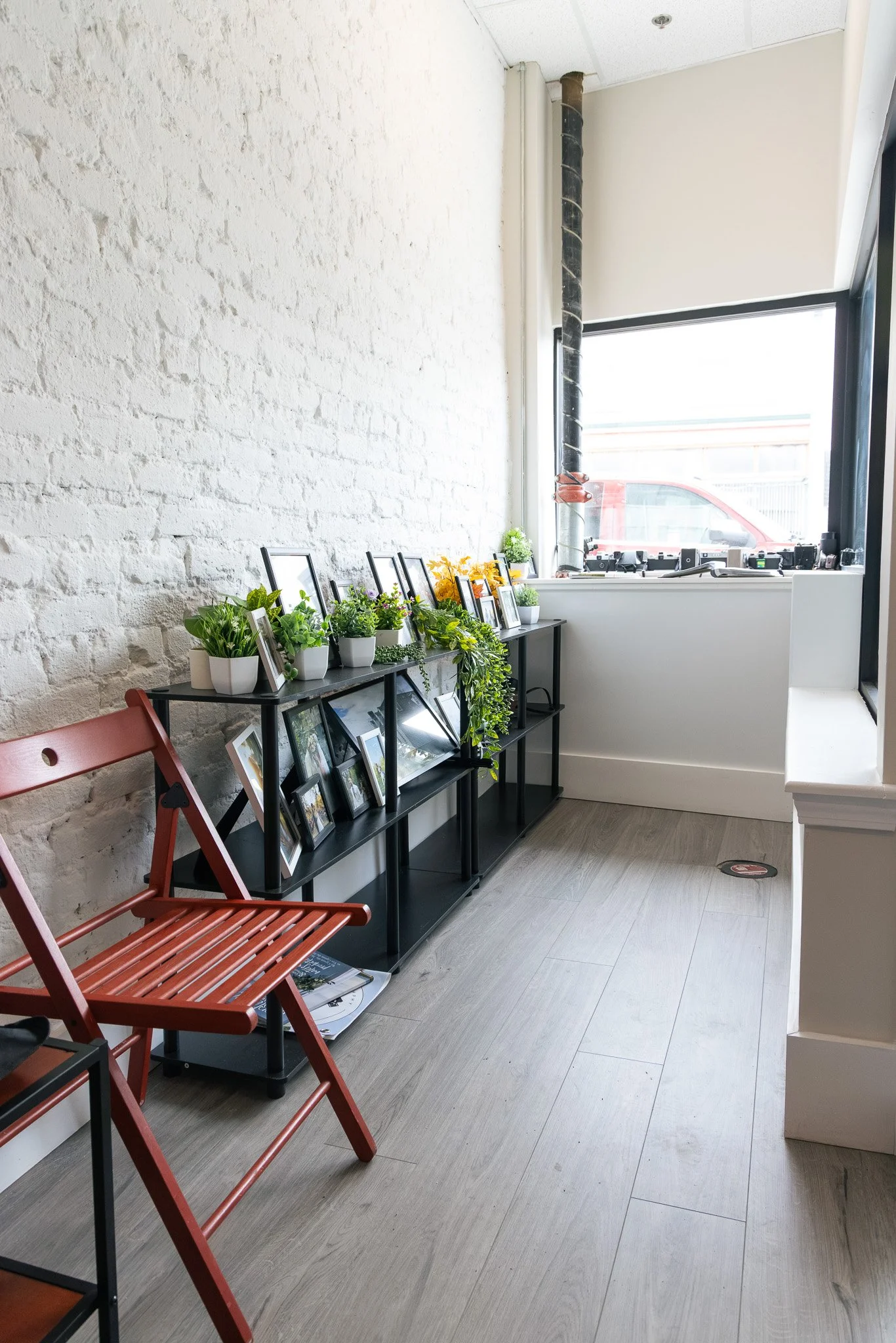 Interior of a modern room featuring a white brick wall, a window with natural daylight, and a black shelf unit decorated with various framed photos and potted plants. A red folding chair is placed in front of the shelf, and a magazine is partially visible on the floor.