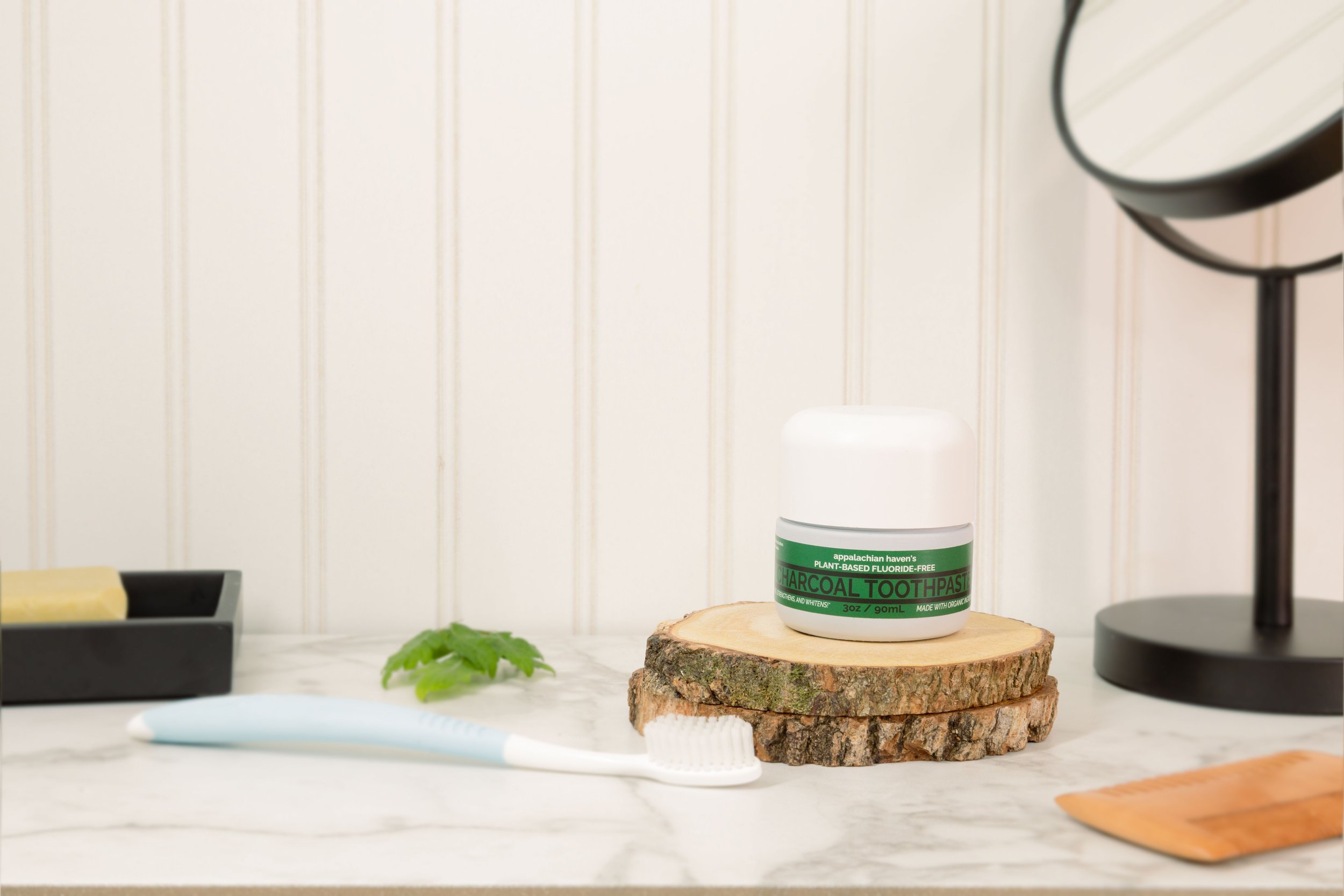 A white jar of Himalayan charcoal toothpaste on a stack of wood slices, set on a white marble surface. Behind the jar, a black mirror and a black container with soap or foam are visible. A green mint leaf and a light blue toothbrush are also on the surface.