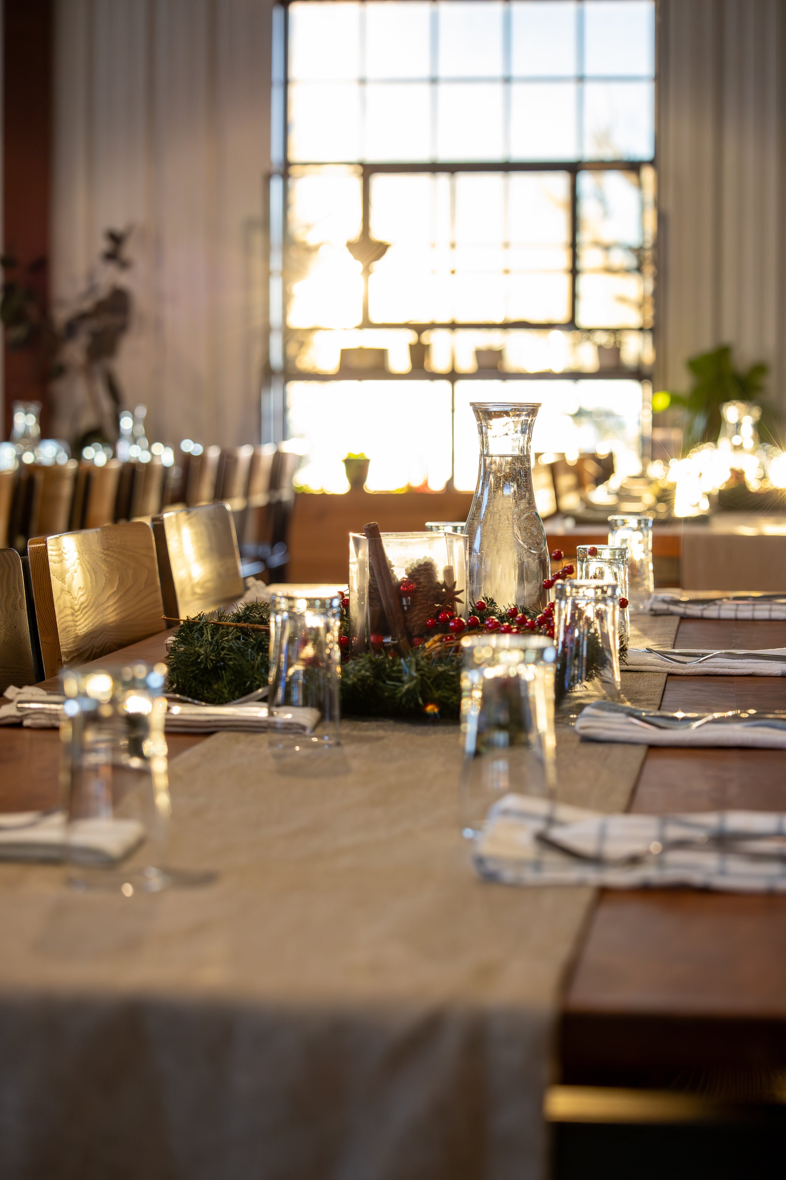 A close-up view of a Christmas table setting with a centerpiece, candles, and glassware, in a well-lit room with large windows and white curtains.