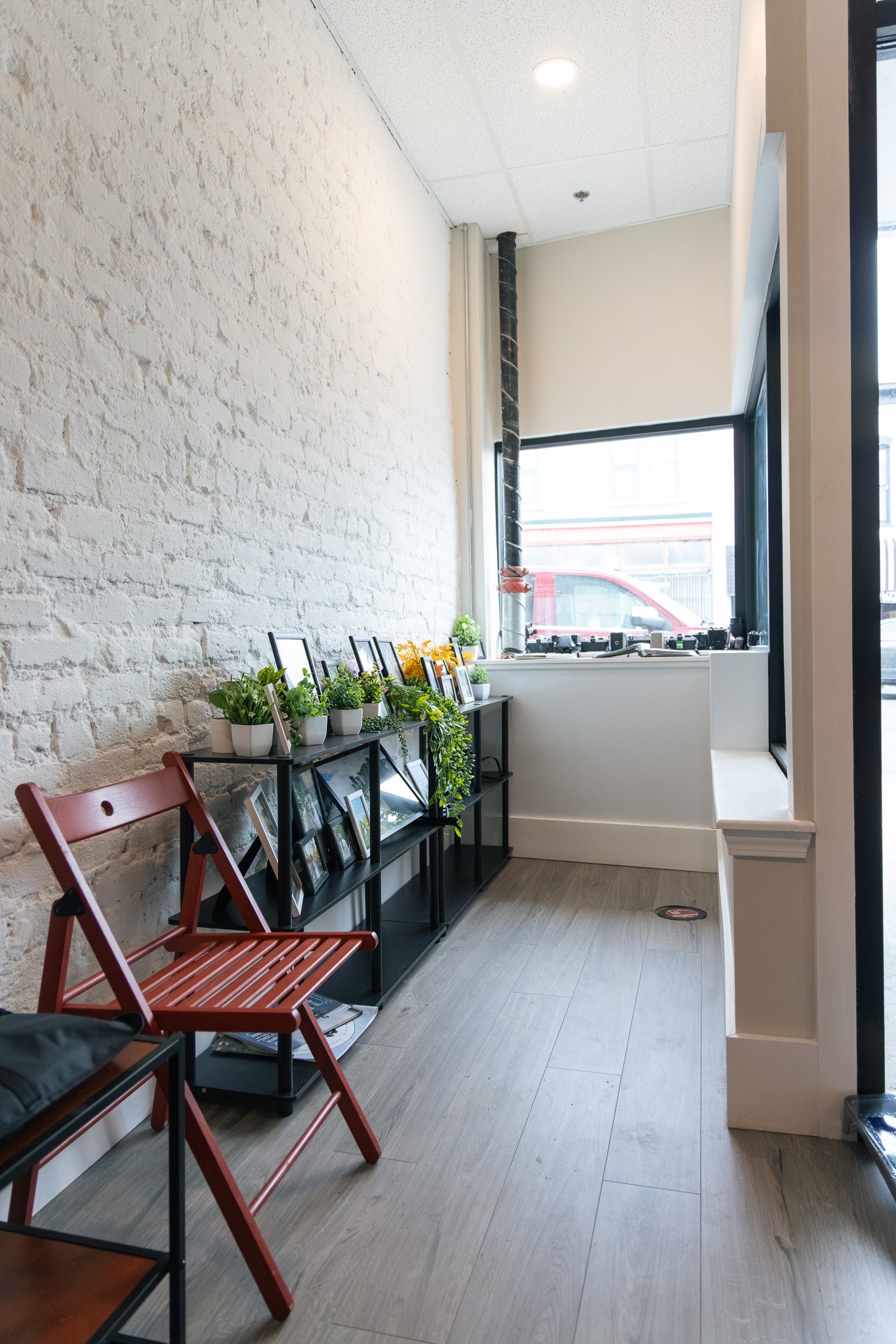 Interior of a cafe or shop with a white brick wall, black metal shelf displaying various framed photos and potted plants, red folding chair, large window letting in natural light, and wooden flooring.
