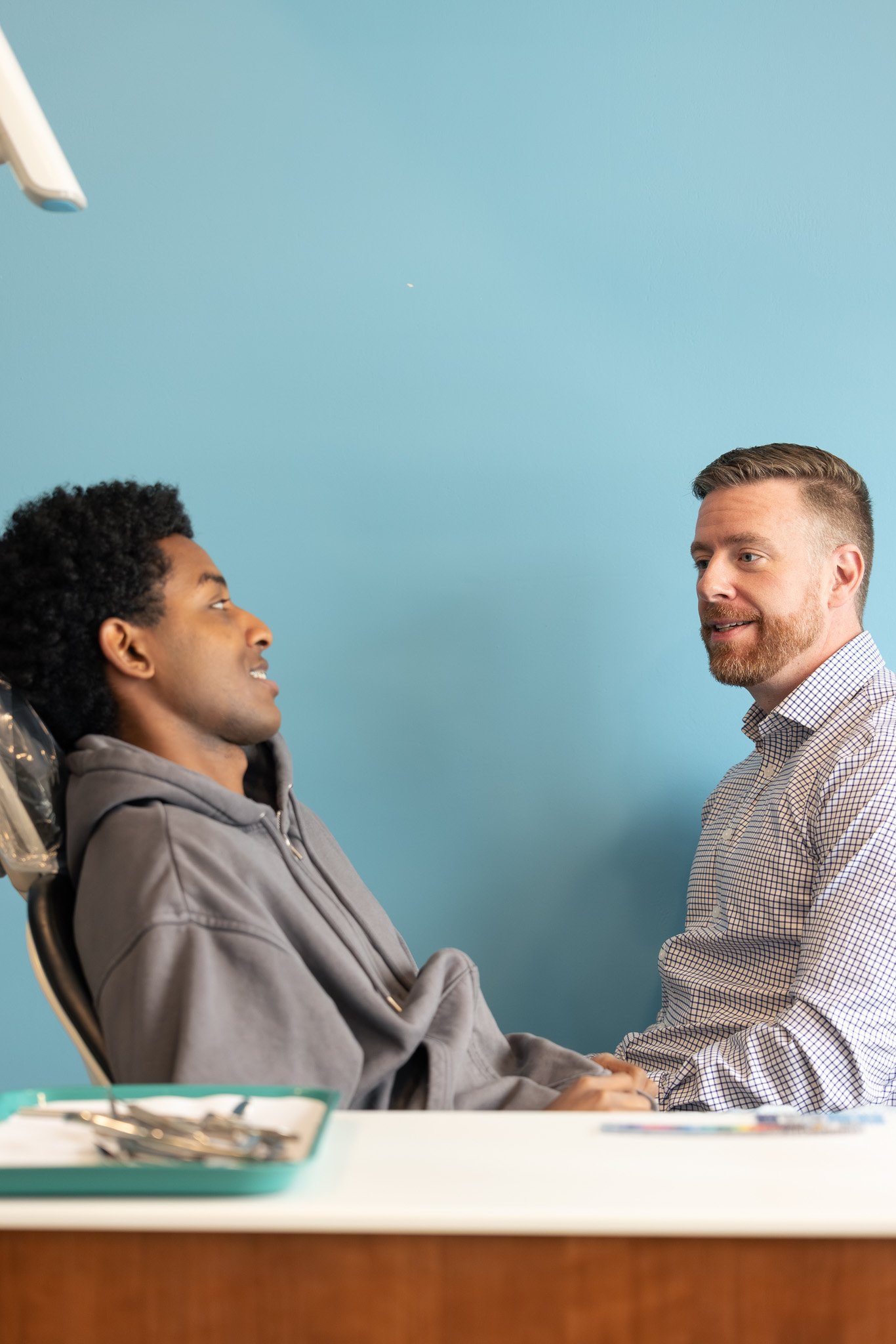 A man with a beard and a checkered shirt holding hands with a young man sitting in a chair, with a dental tray in the foreground and a light blue wall in the background.