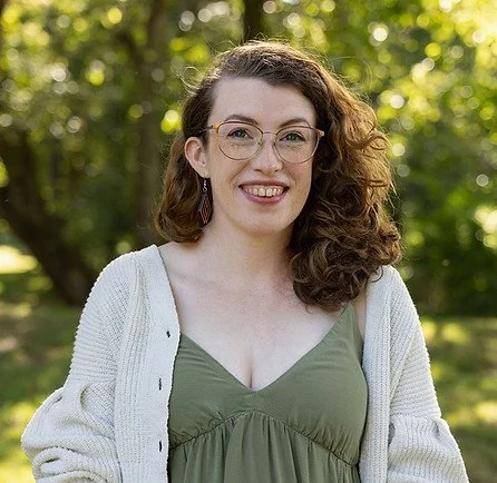 A woman with curly brown hair, glasses, and earrings, smiling outdoors with trees and greenery in the background.