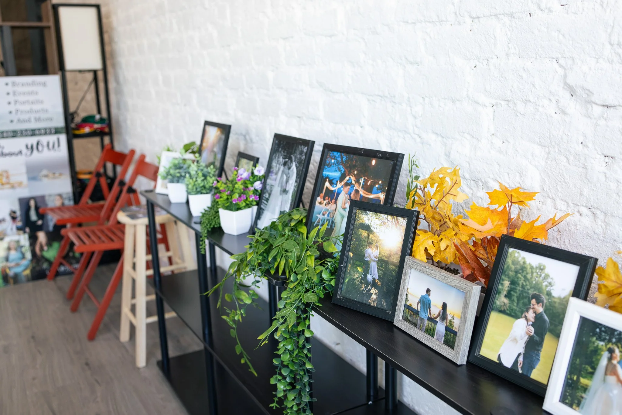 Display of framed photographs and potted plants against a white brick wall, with red chairs and a sign in the background.