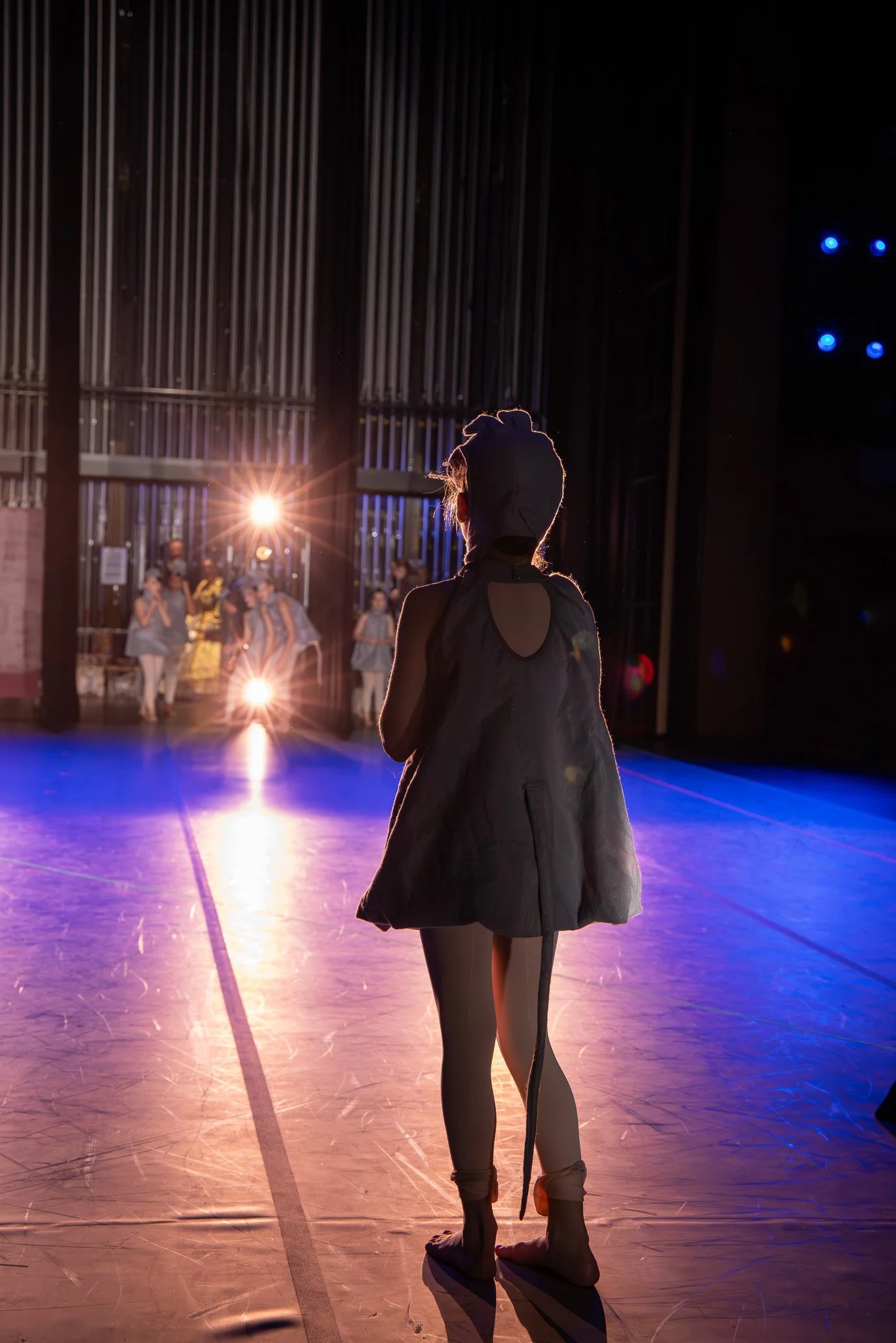 A young female ballet dancer with a bun hairstyle standing on a dance floor in a studio, backlit by stage lights, with a group of dancers in the background.