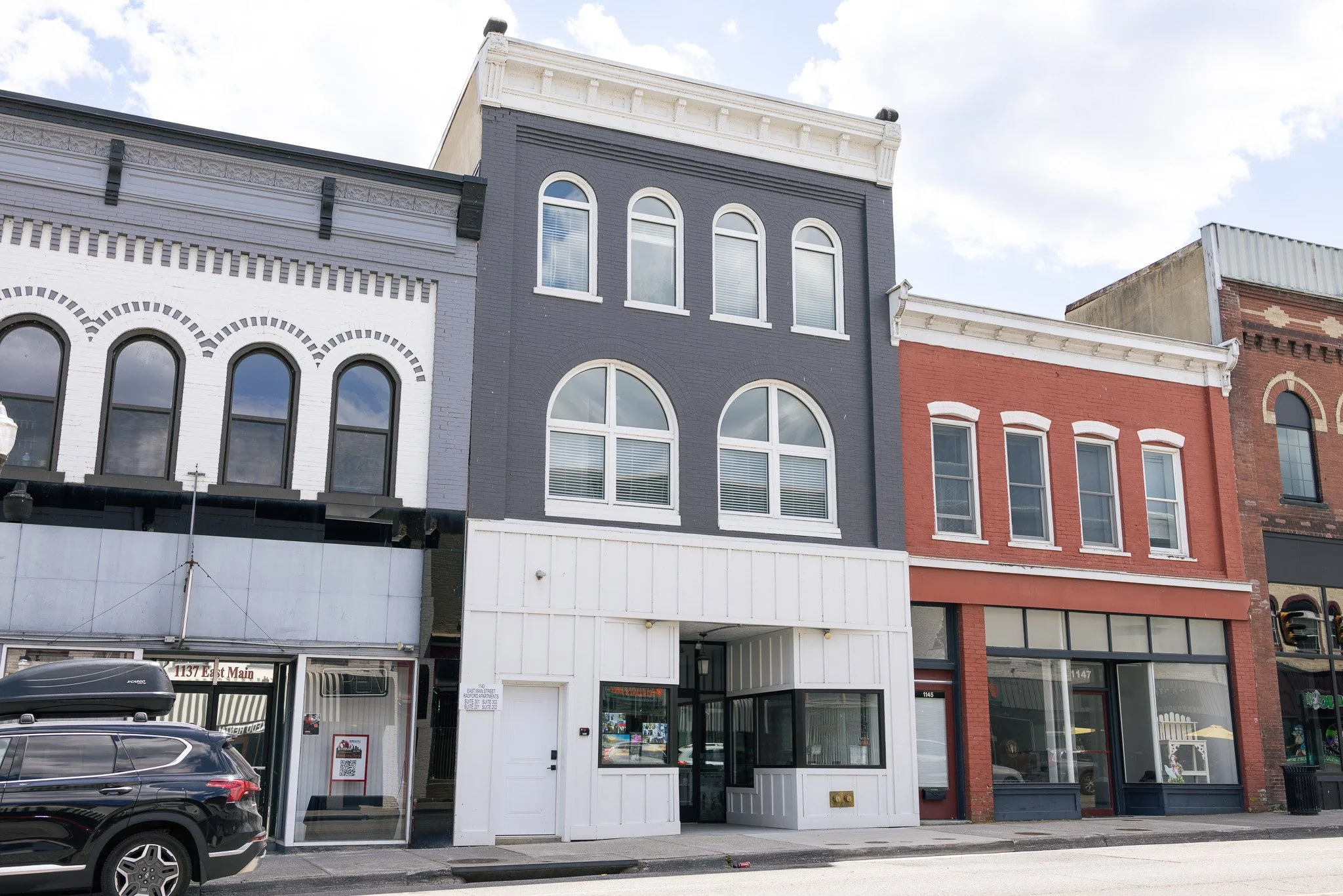 A row of storefront buildings with different architectural styles and colors, including white, gray, and red brick, under a partly cloudy sky. There are cars parked in front of the buildings.