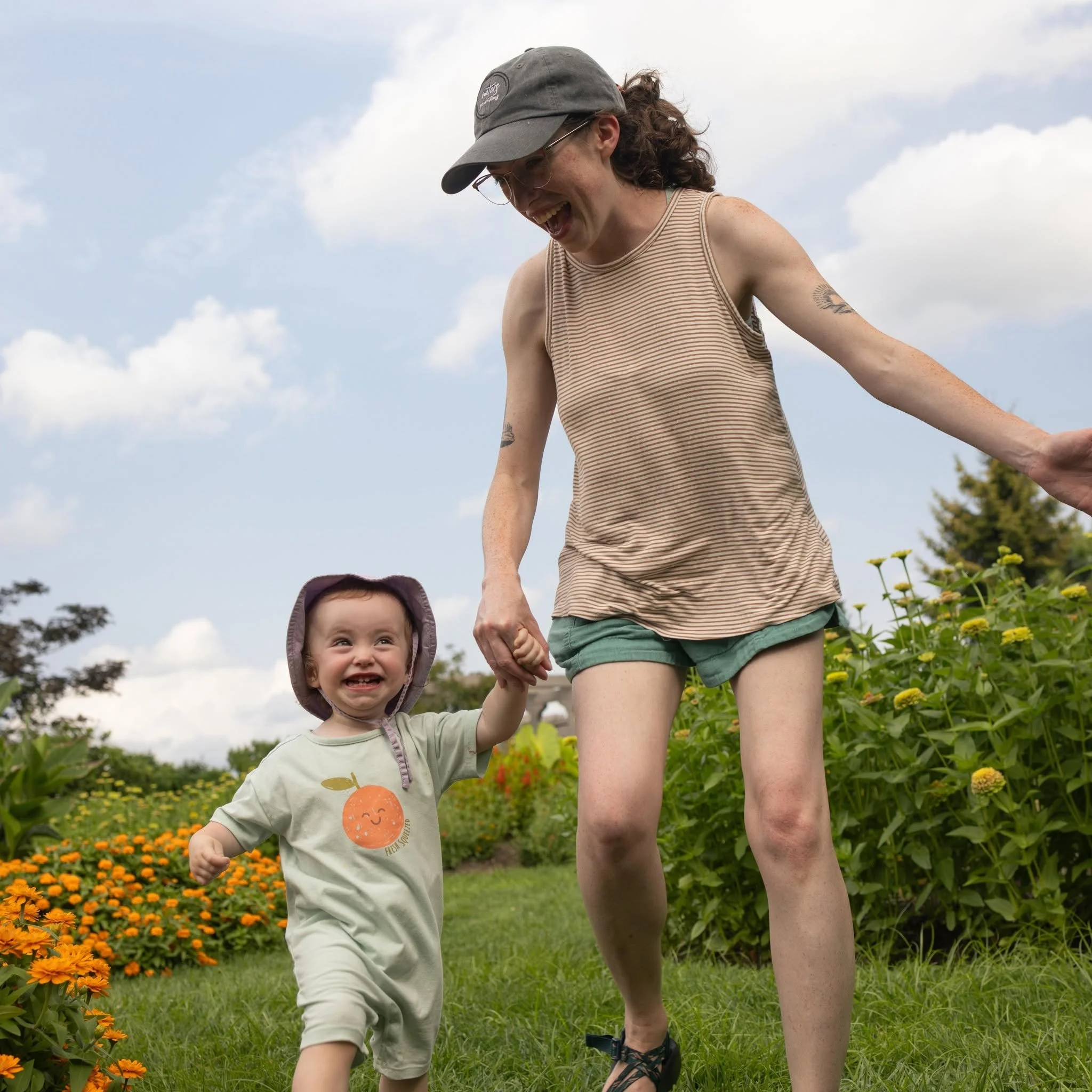 A woman and a young girl are outdoors in a flower garden, holding hands and smiling. The woman is wearing a striped tank top, shorts, sandals, glasses, and a cap, while the girl is wearing a light-colored shirt with a smiley face and a sun graphic, and a hood on her head. The sky is partly cloudy and there are vibrant orange flowers around them.