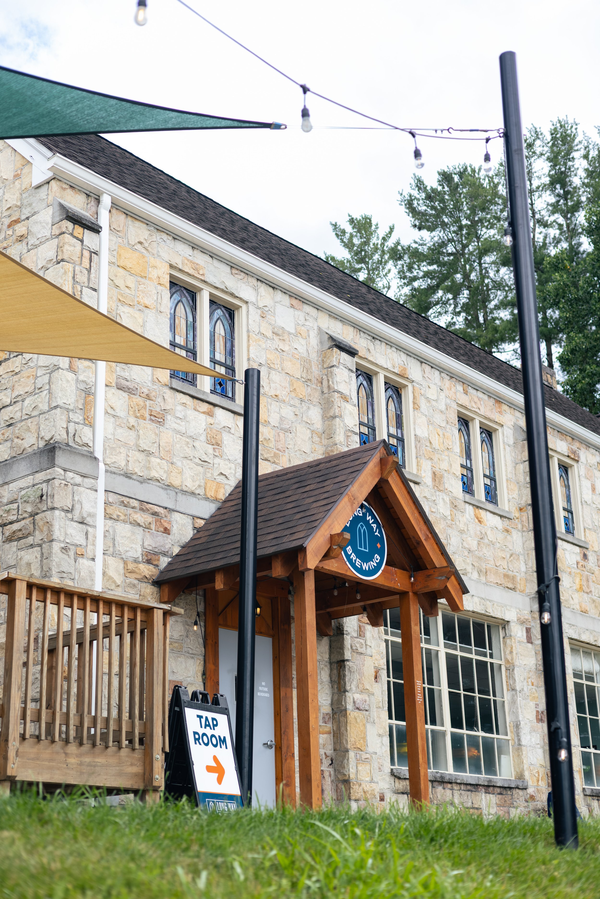 The image shows the exterior of a stone building with stained glass windows. In front, there is a wooden entrance with a sign directing to a tap room. There is also a black sign with an orange arrow pointing to the right that says "Tap Room." String lights are hanging overhead, and a green shade canopy is partially visible at the top left. Some trees are visible in the background against a cloudy sky.