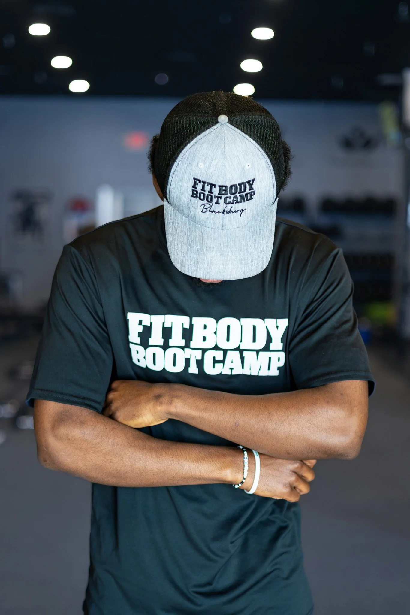Person wearing a black and gray baseball cap with the words "FITBODY BOOTCAMP Blackburn" embroidered on it, standing with arms crossed in a gym.