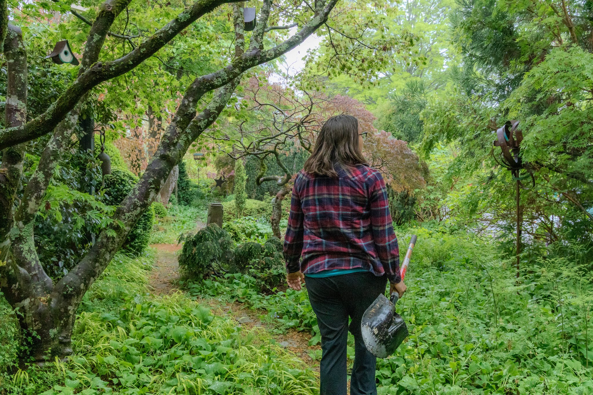 A person with glasses and long dark hair walking in a lush, green garden holding a small shovel and a black container.