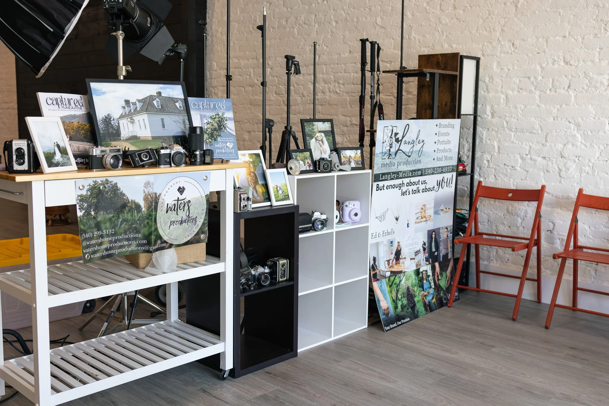 A photography studio display with cameras, framed photos, and promotional posters, set against a white brick wall and hardwood floor, with red chairs to the right.