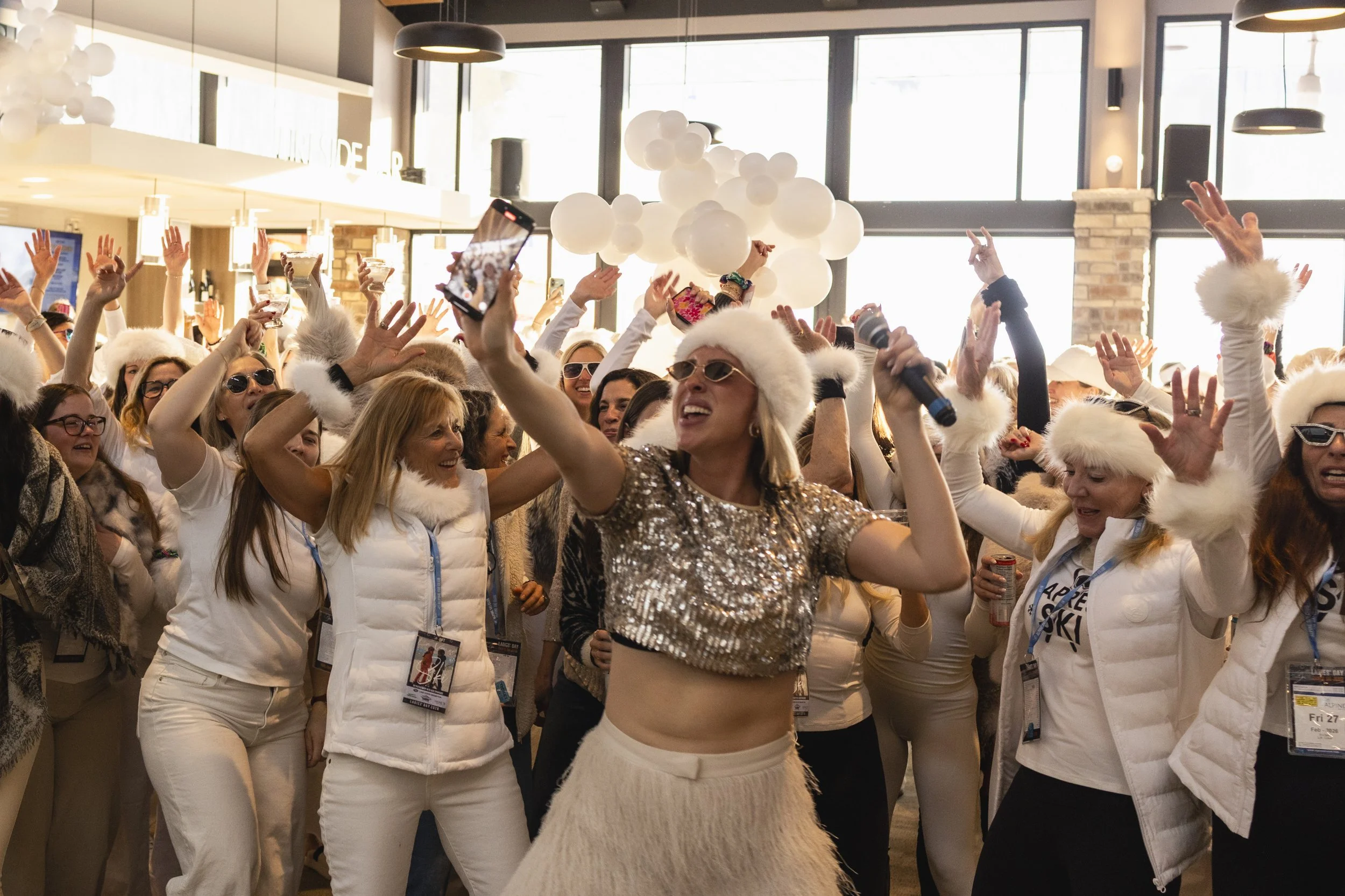 Rachel Molenda wearing sparkly silver top and white tassel pants on a microphone MC-ing at Alpine Ski Resort event with women dancing in the crowd