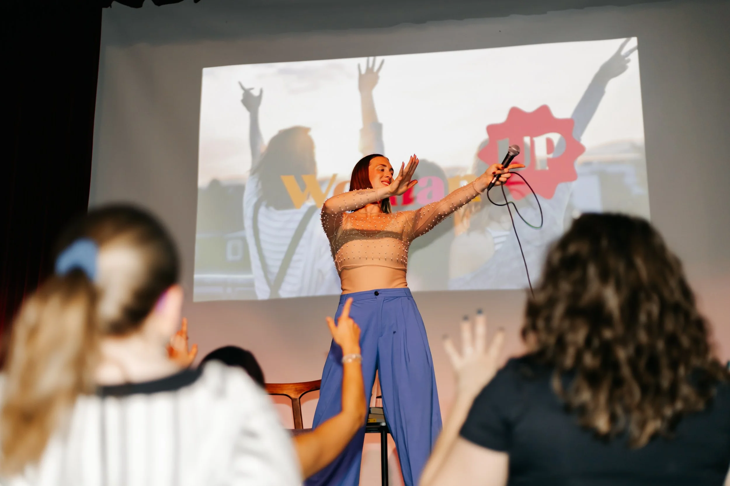 Rachel Molenda wearing blue pants and beige top on a microphone MC-ing at brand activation with women dancing in the audience