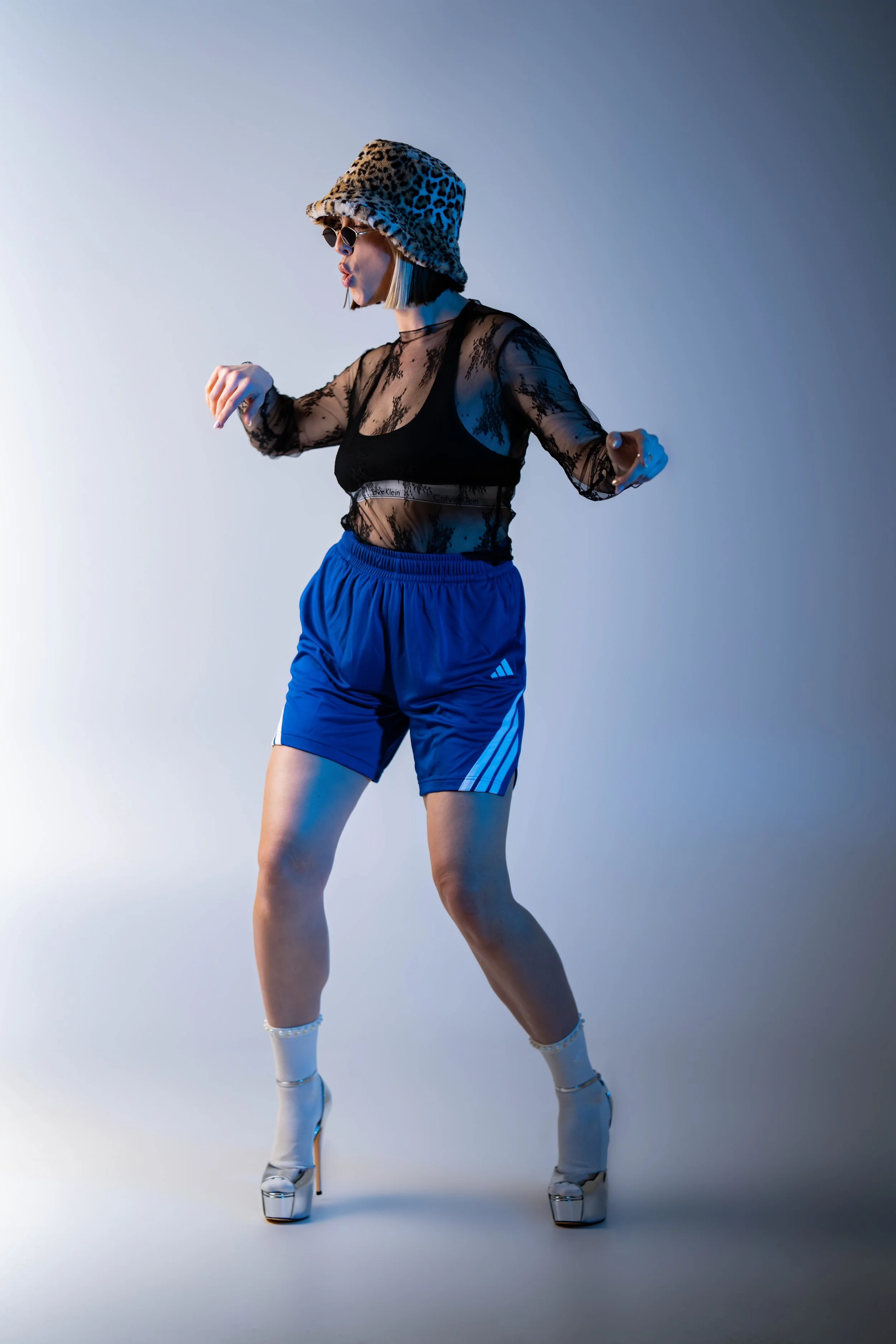Rachel wearing a leopard print fuzzy bucket hat, a lace black top, blue adidas shorts and raver sunglasses dancing