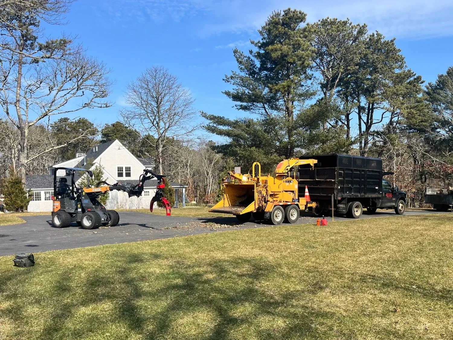 A residential yard with a black mini skid steer loader, a yellow wood chipper, and a black work truck. The yard has grass, a paved area, and trees with a house in the background.