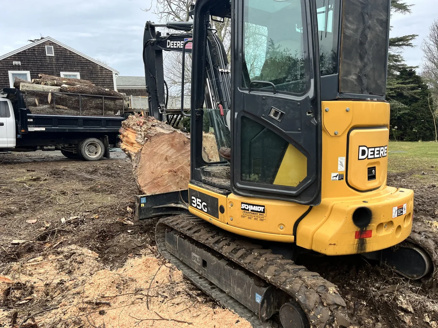 A yellow Deere compact excavator (model 35G) is digging wood debris, lifting a large log at a construction site or yard, with a truck loaded with logs in the background and a house and trees in the distance.