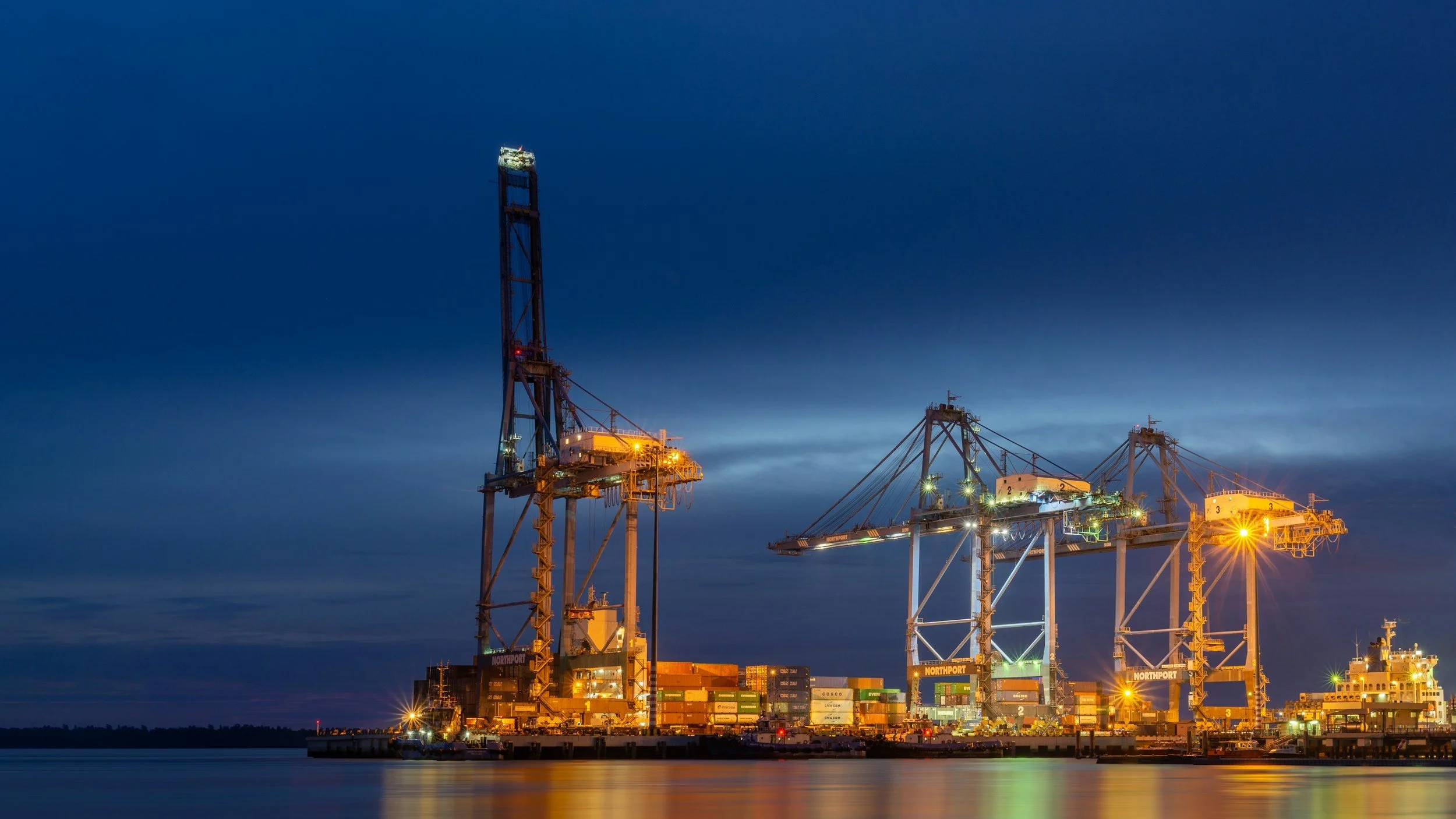 Nighttime view of several large offshore oil drilling rigs with illuminated structures and stacked shipping containers, reflecting in calm water.