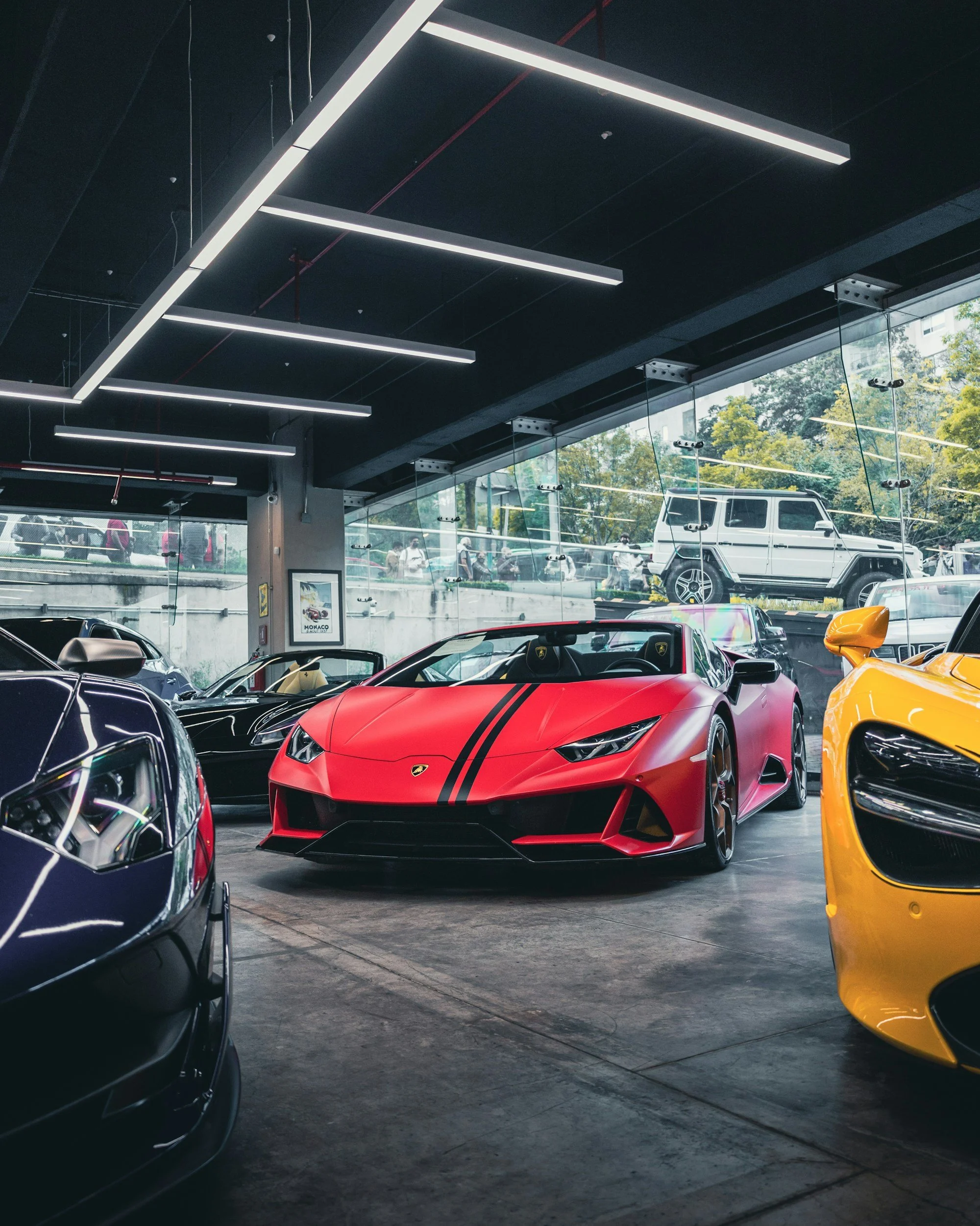 View of luxury cars inside a modern showroom with large windows, including a red Lamborghini, a black car, and part of a yellow sports car in the foreground, with reflections on the dark polished floor and sleek ceiling lights.