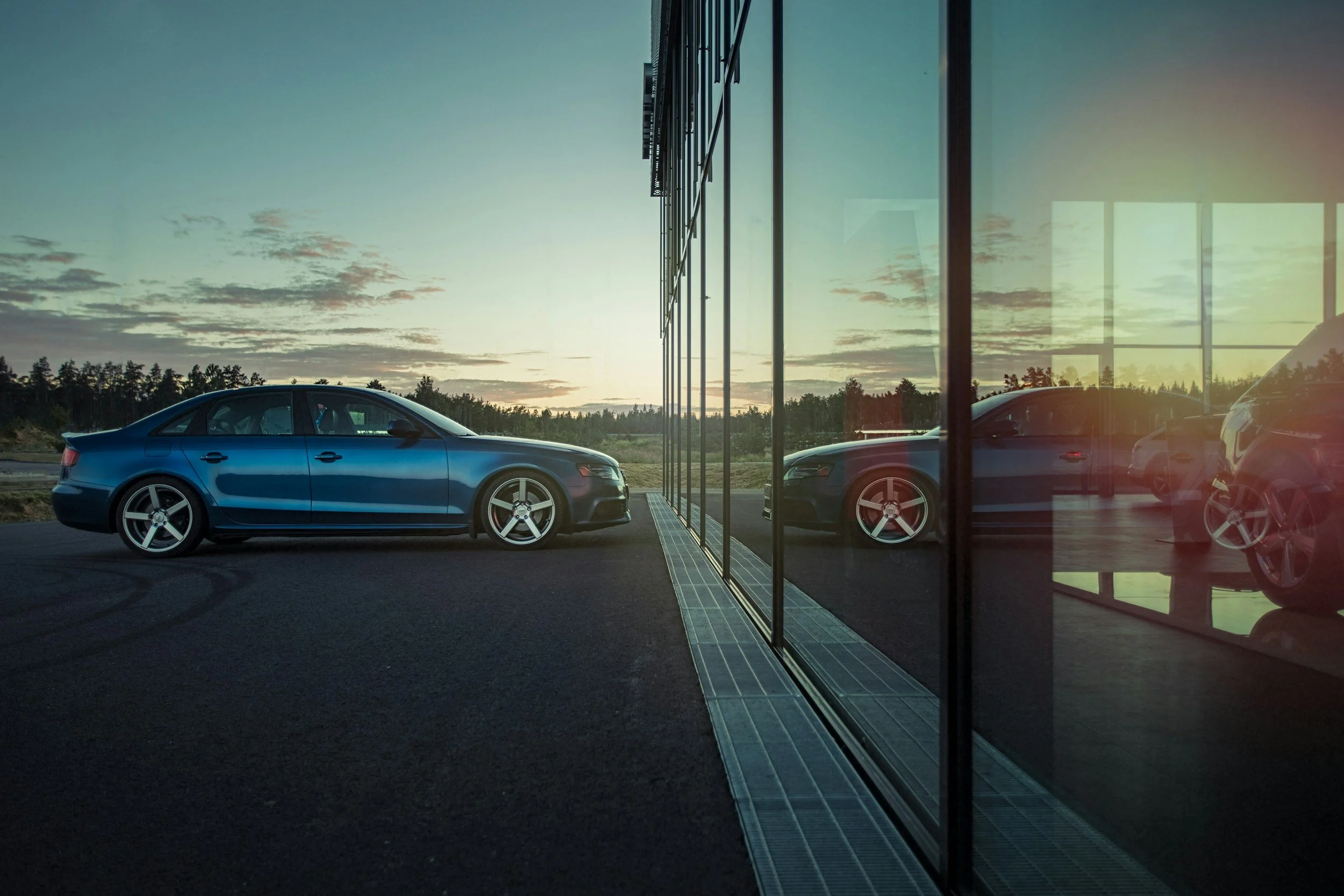 Side view of a blue modern sedan parked outside of a building with large glass windows, reflecting the car and the sky at sunset.
