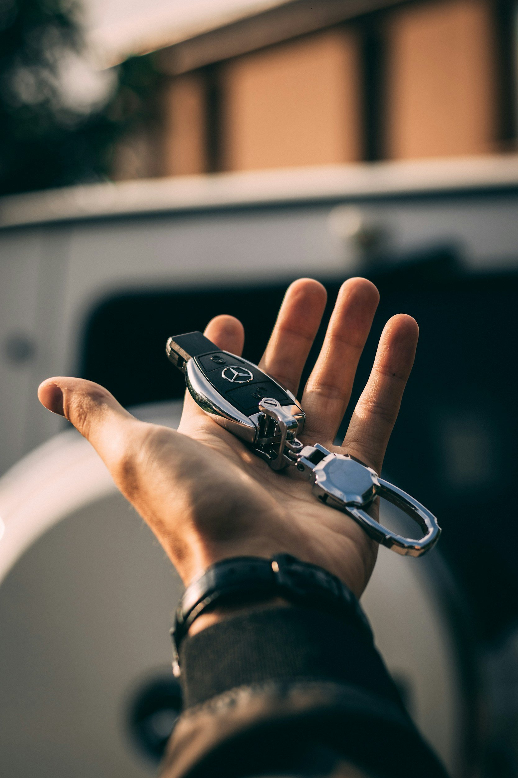 Close-up of a hand holding a Mercedes-Benz car key with a silver chain, against a blurred background.