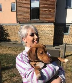 Smiling woman with short gray hair holding a brown dog outdoors on a sunny day.