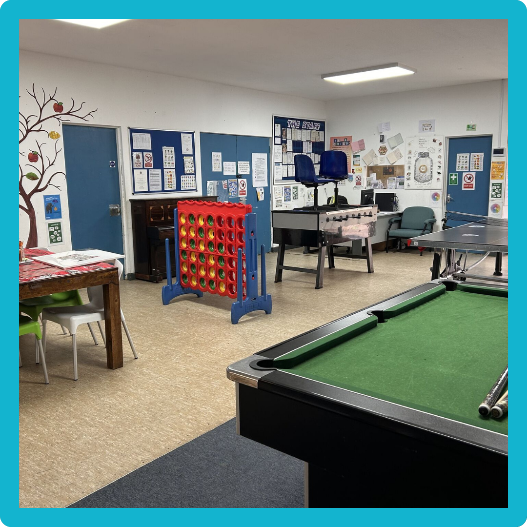A recreation room with a pool table, a Connect Four game, a foosball table, a computer station, and chairs. There are bulletin boards and posters on the walls, and a painted tree with apples on the left.
