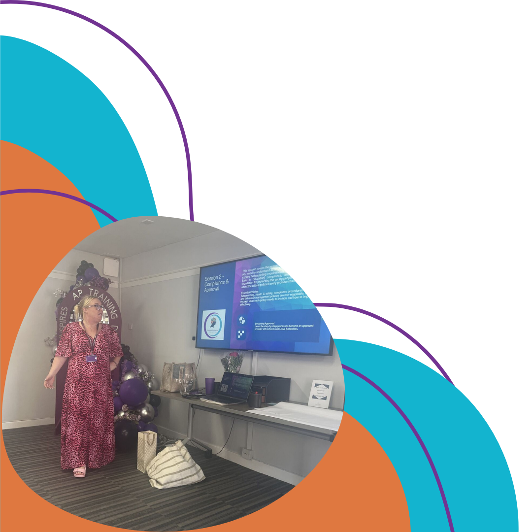 A woman in a pink and black leopard print dress stands in a training or conference room next to a large monitor displaying a presentation slide. The room has decorations including purple and silver balloons and a banner in the background. There is a table with electronic equipment and bags on the floor.