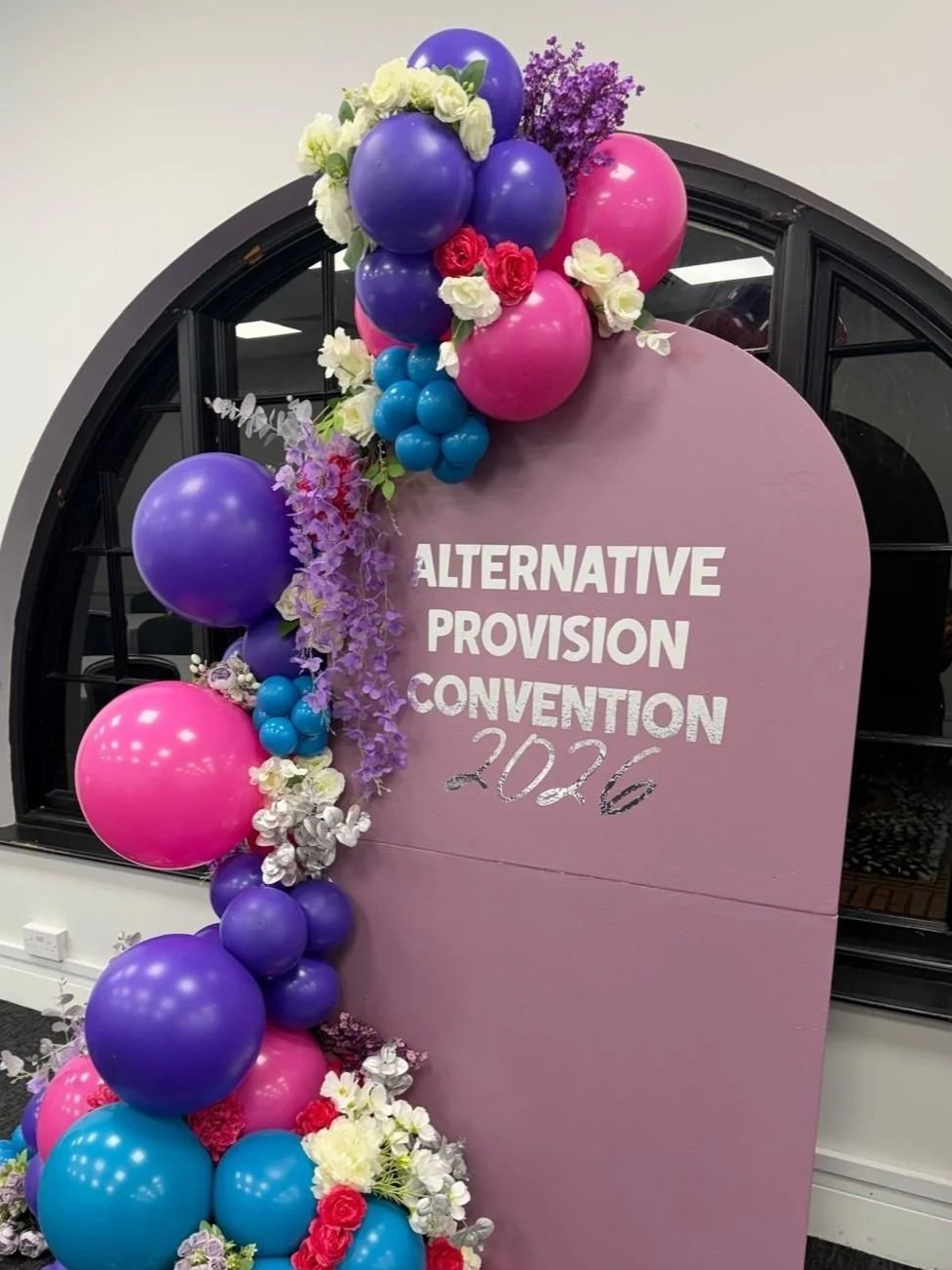 Decorative display with colorful balloons and flowers surrounding a pink sign that reads 'ALTERNATIVE PROVISION CONVENTION 2026'.