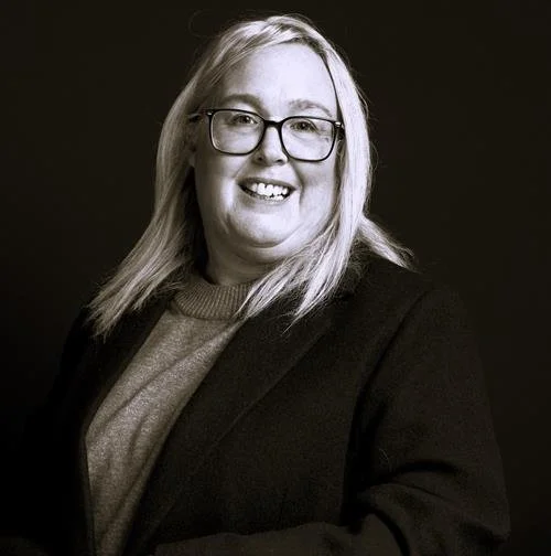 A woman with long blonde hair, wearing glasses, a gray sweater, and a dark blazer, smiling in a professional portrait with a dark background.