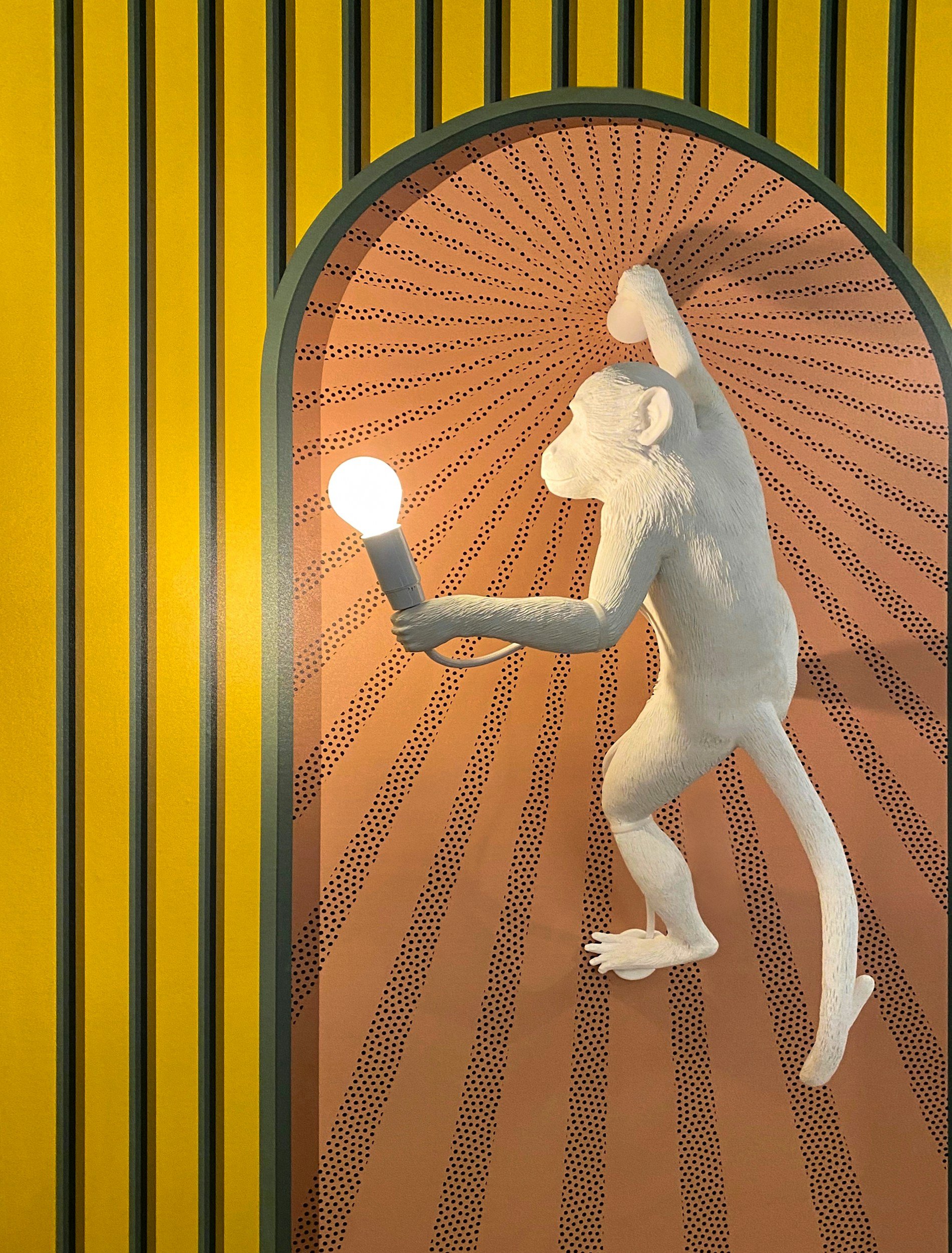 A white sculpture of a baboon holding a lightbulb, set against a colorful background with yellow, black, and green vertical stripes and a radial pattern behind the sculpture.