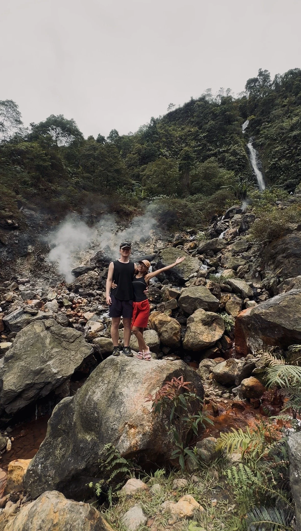 Two people standing on large rocks in a rocky outdoor area with a waterfall cascading down a hillside in the background, surrounded by trees and vegetation.